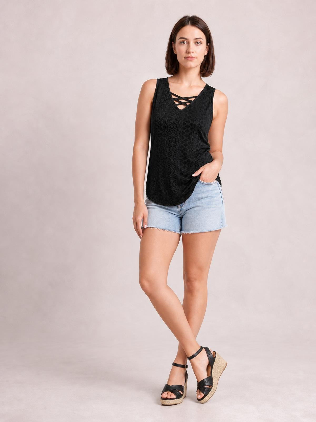 Model poses in a black eyelet wide strap tank with a relaxed silhouette, rounded hem, and crisscross V-neck detail, paired with denim shorts.