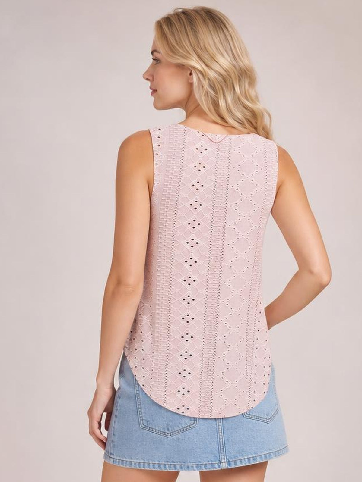 A model poses in a pale pink eyelet tank with wide straps, a relaxed straight silhouette, rounded hem, and intricate vertical cutout patterns visible from the back.