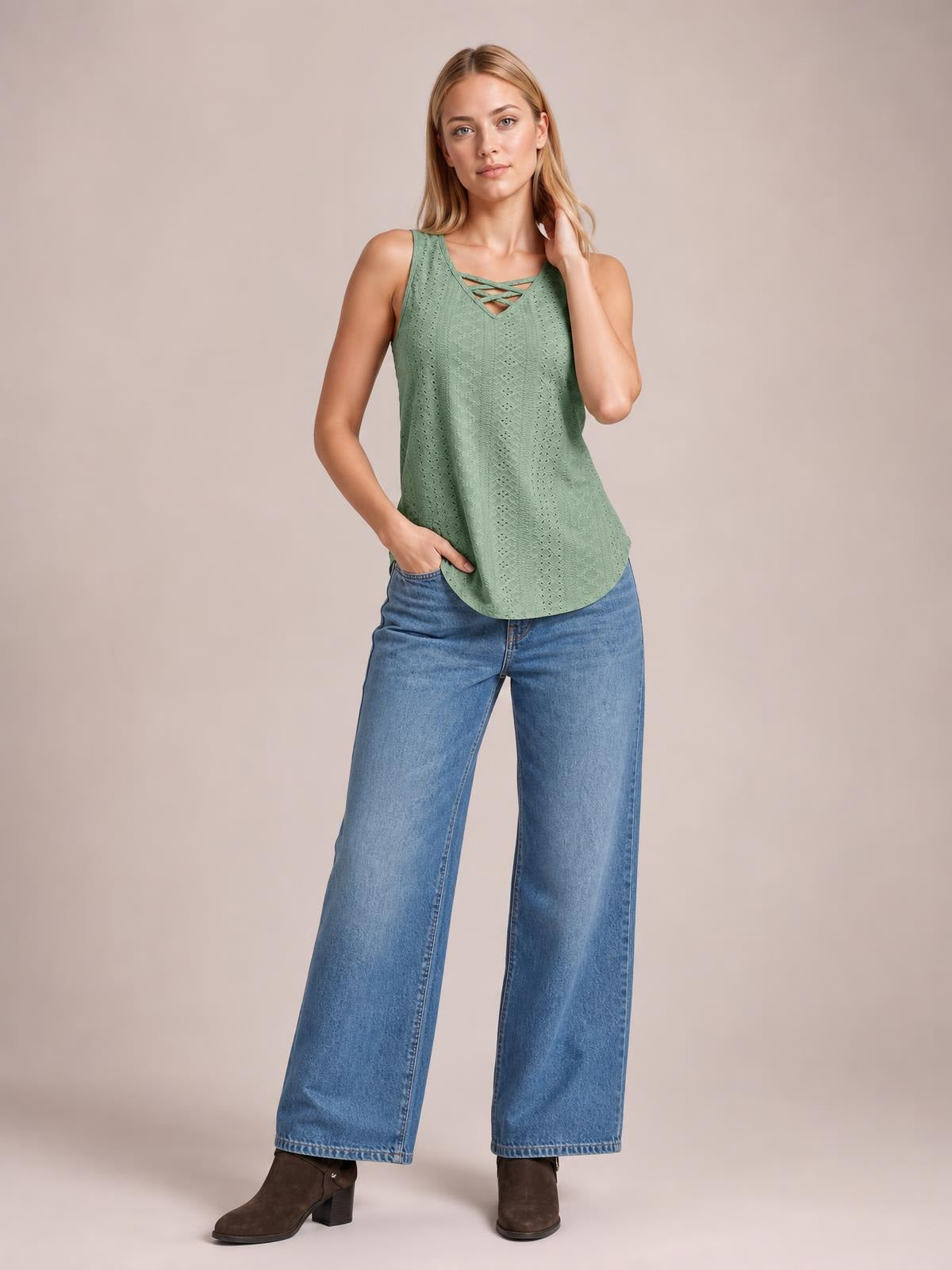 A model wears a sage green eyelet wide-strap tank with a relaxed silhouette, crisscross neckline detail, and curved hem while posing with one hand in her pocket.