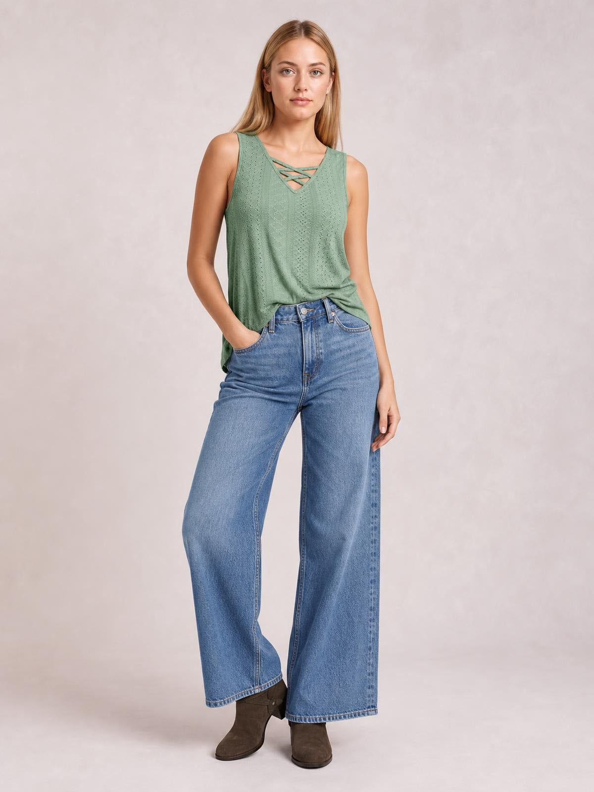 A model wears a sage green eyelet wide strap tank with a relaxed silhouette, V-neck crisscross detail, and gently draped construction, posing with one hand in her jeans pocket.