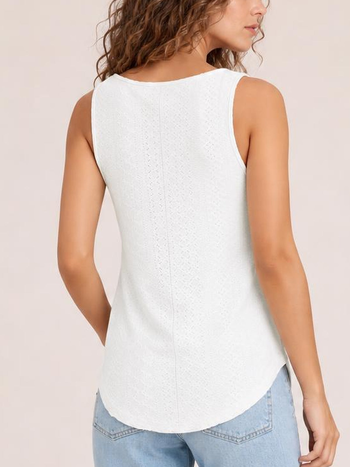 A model poses with one hand at her hip, showing the white eyelet tank’s wide straps, opaque relaxed silhouette, and curved high-low hem.