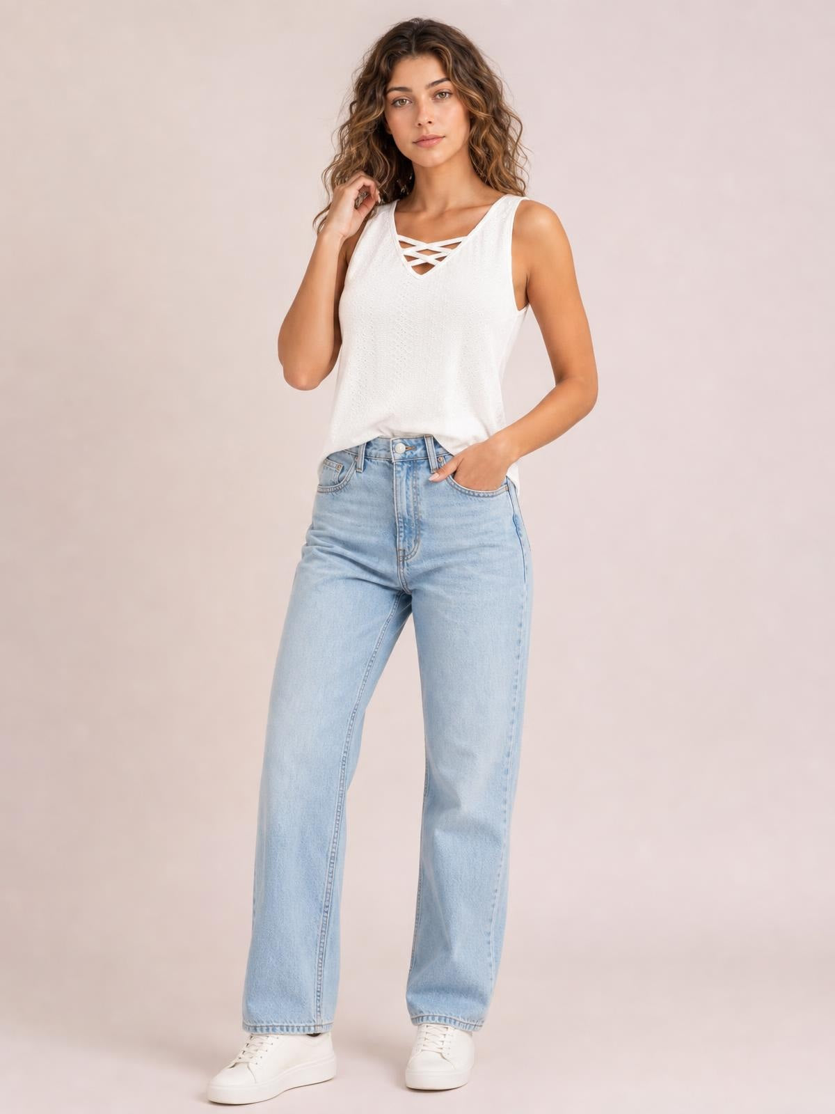 A model poses in a white opaque eyelet tank with wide straps, a relaxed silhouette, and a crisscross neckline detail, one hand in her jeans pocket.