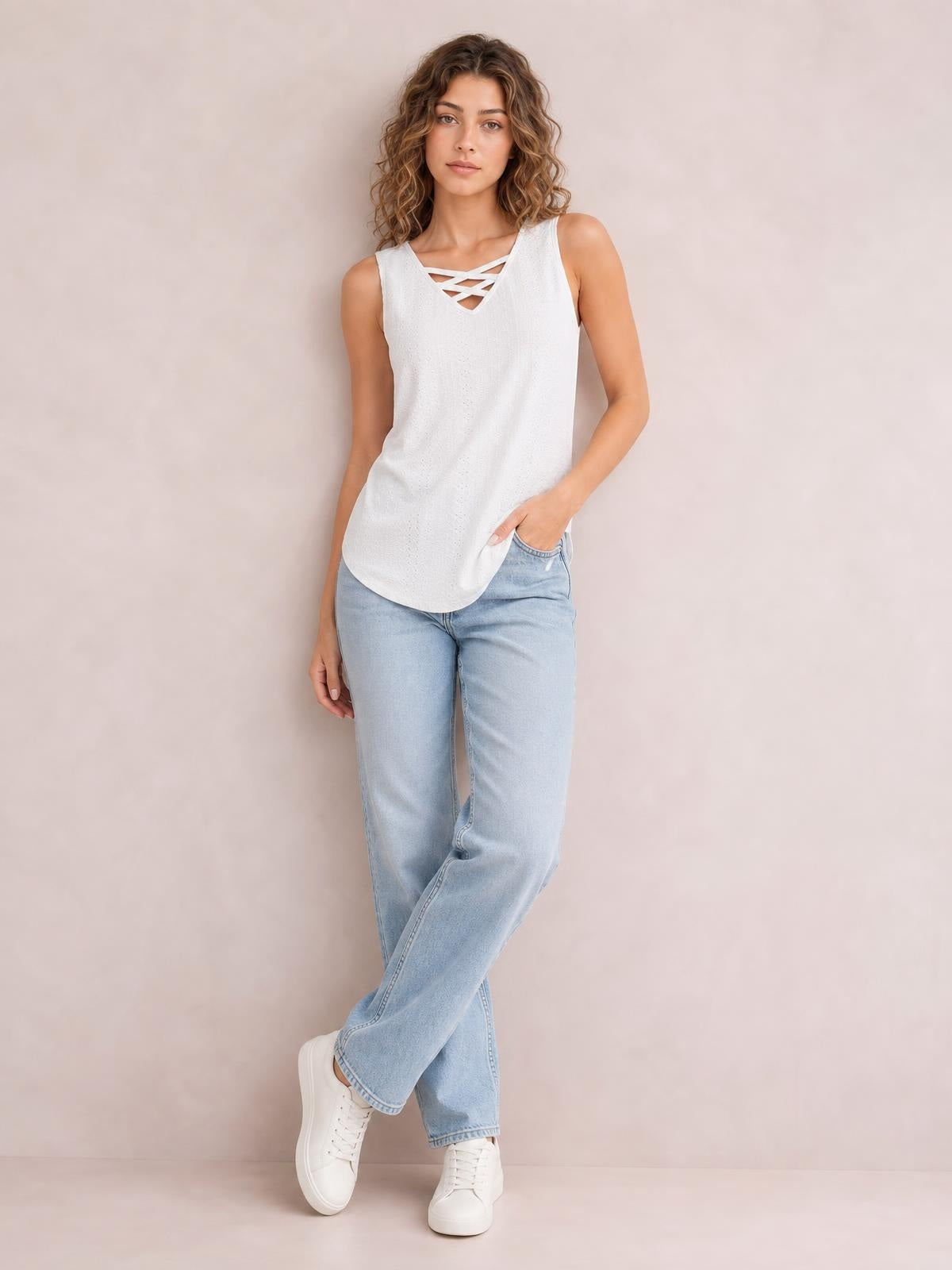 Model wears a white eyelet wide-strap tank with a relaxed silhouette, rounded hem, and crisscross neckline, posed with one hand in her jeans pocket.