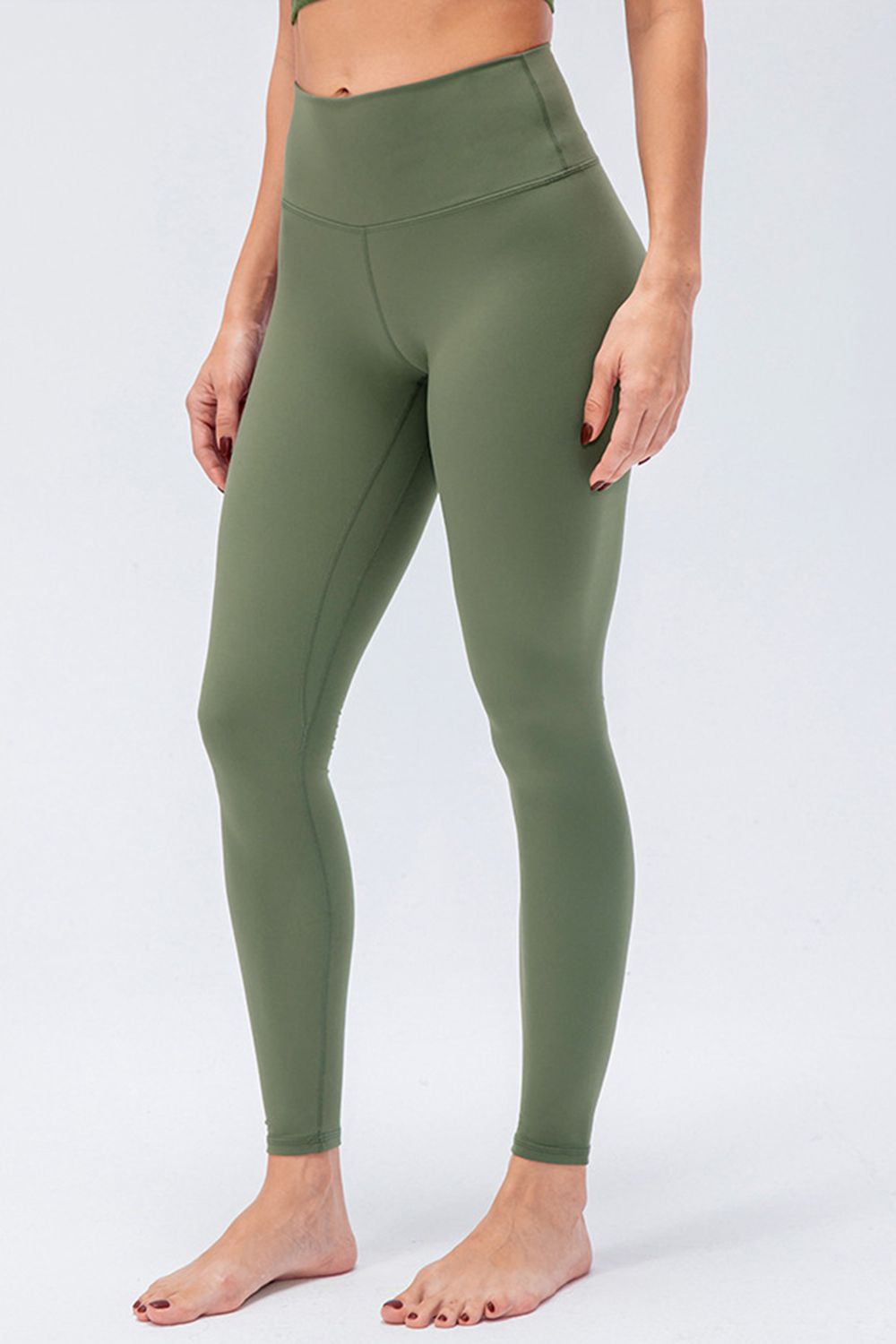 Model wears olive green wide-waistband slim-fit active leggings with a high-rise silhouette, ankle length, and visible curved seam construction.