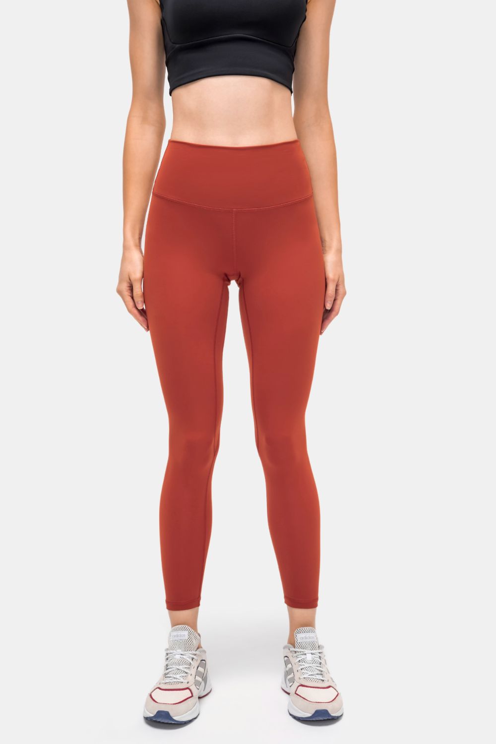 Model stands facing forward in rust-red high-rise sports leggings with a slim, ankle-length silhouette, wide waistband, and visible front seam construction.
