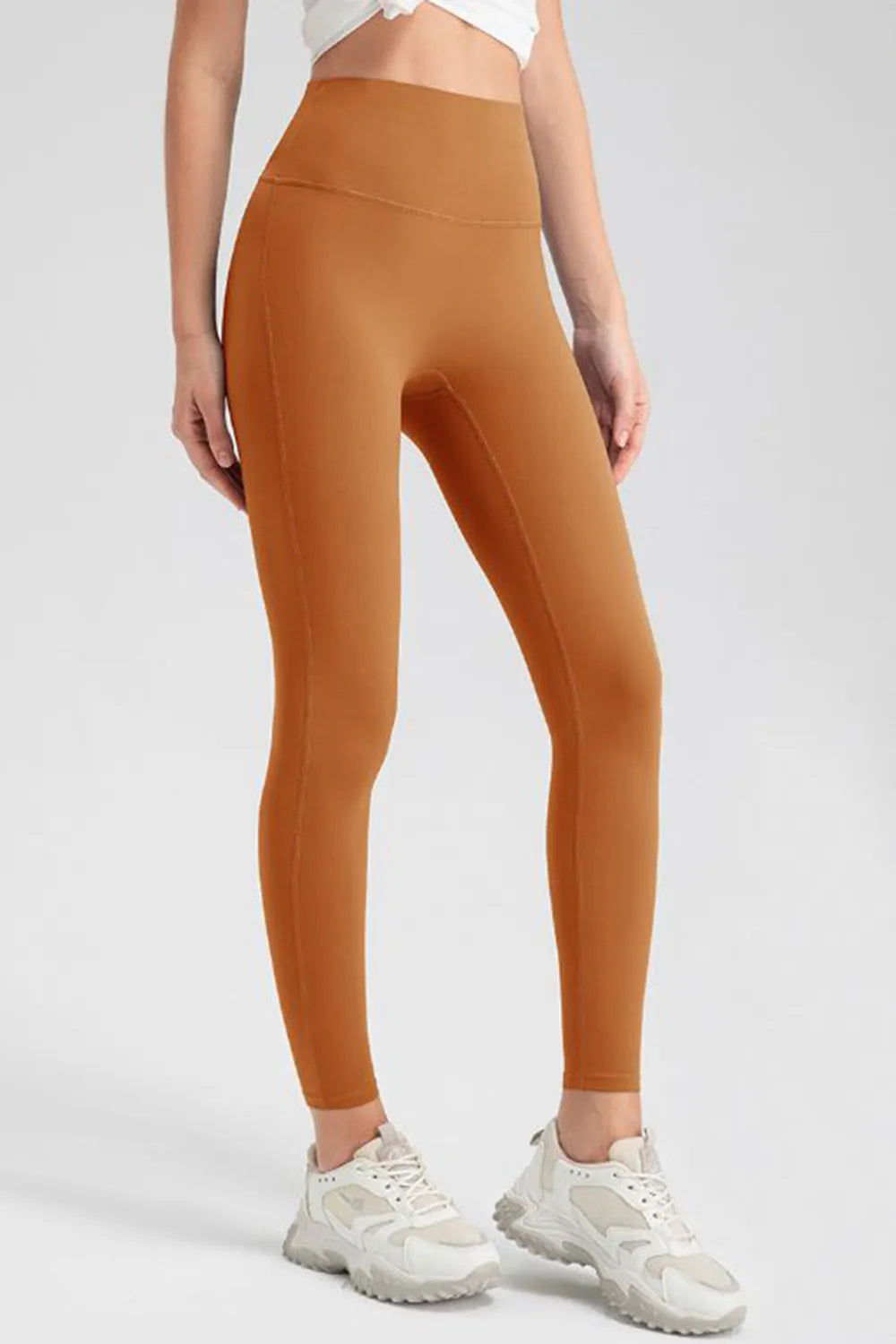 Model wears rust-orange high-waist skinny active pants with a sleek ankle-length silhouette, stretchy fit, and visible flat seams while standing with legs slightly apart.