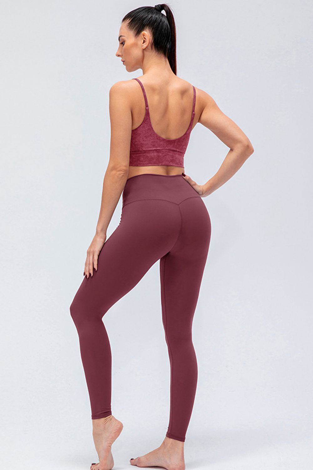 Model poses in burgundy wide-waistband slim-fit active leggings with a high-rise silhouette, smooth construction, and full-length tapered legs.