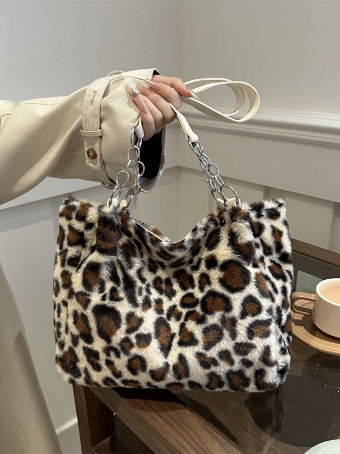 A large faux fur leopard shoulder bag features a softly rounded rectangular silhouette, plush cream, brown, and black print, and chain-linked strap attachments.