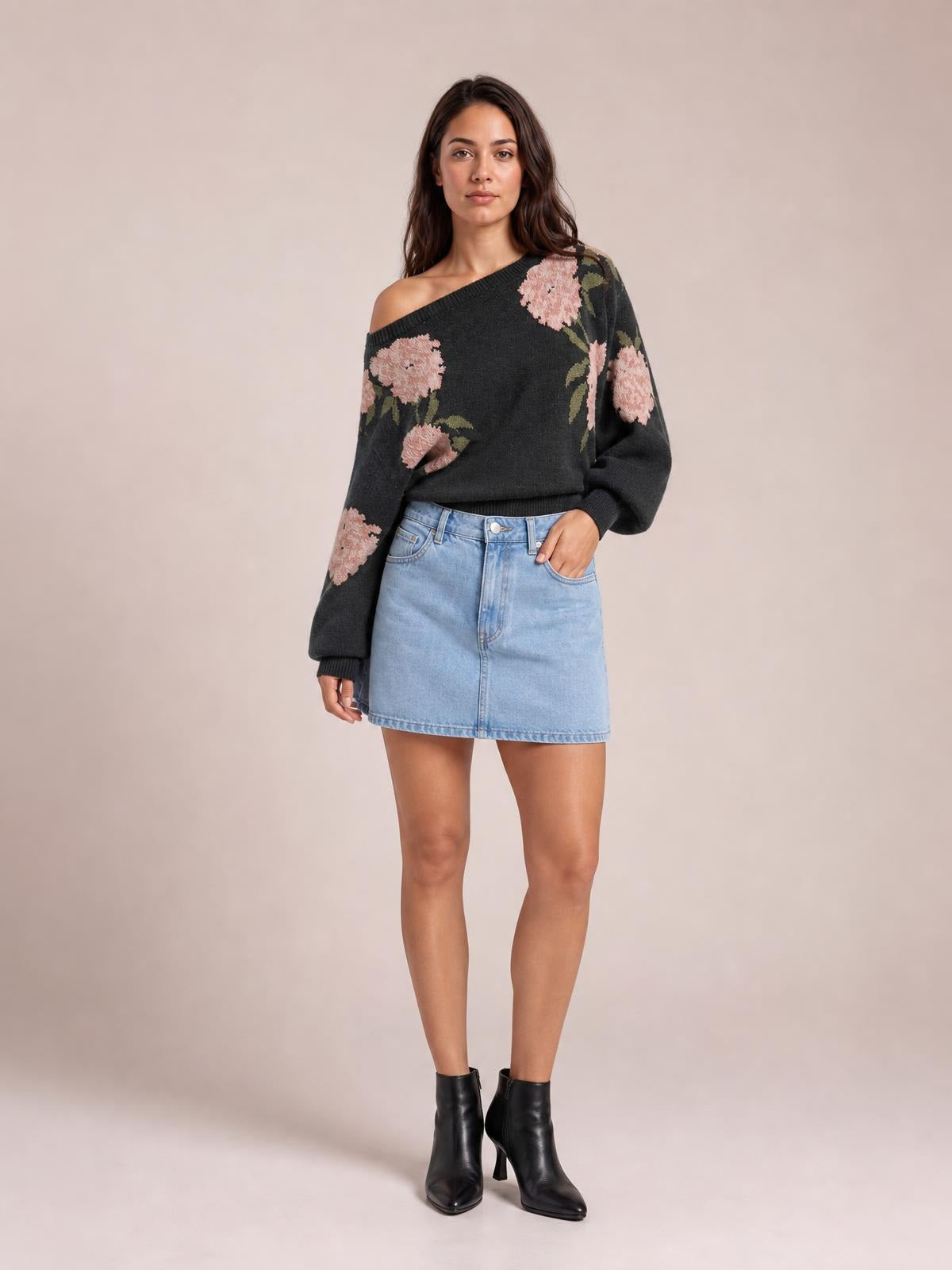 A model wears a charcoal floral boat-neck long-sleeve sweater with pink blooms, ribbed cuffs and hem, and a relaxed silhouette, posing with one hand in her pocket.
