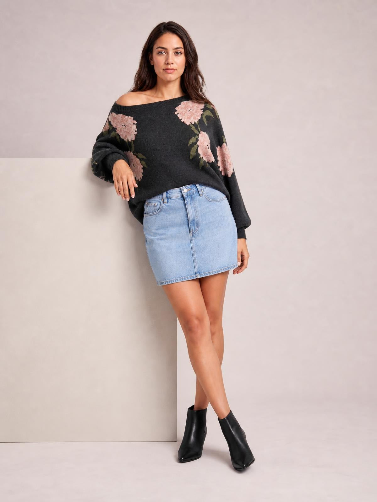 Model leans against a surface wearing a relaxed black floral boat neck long sleeve sweater with pink flower and green leaf accents.