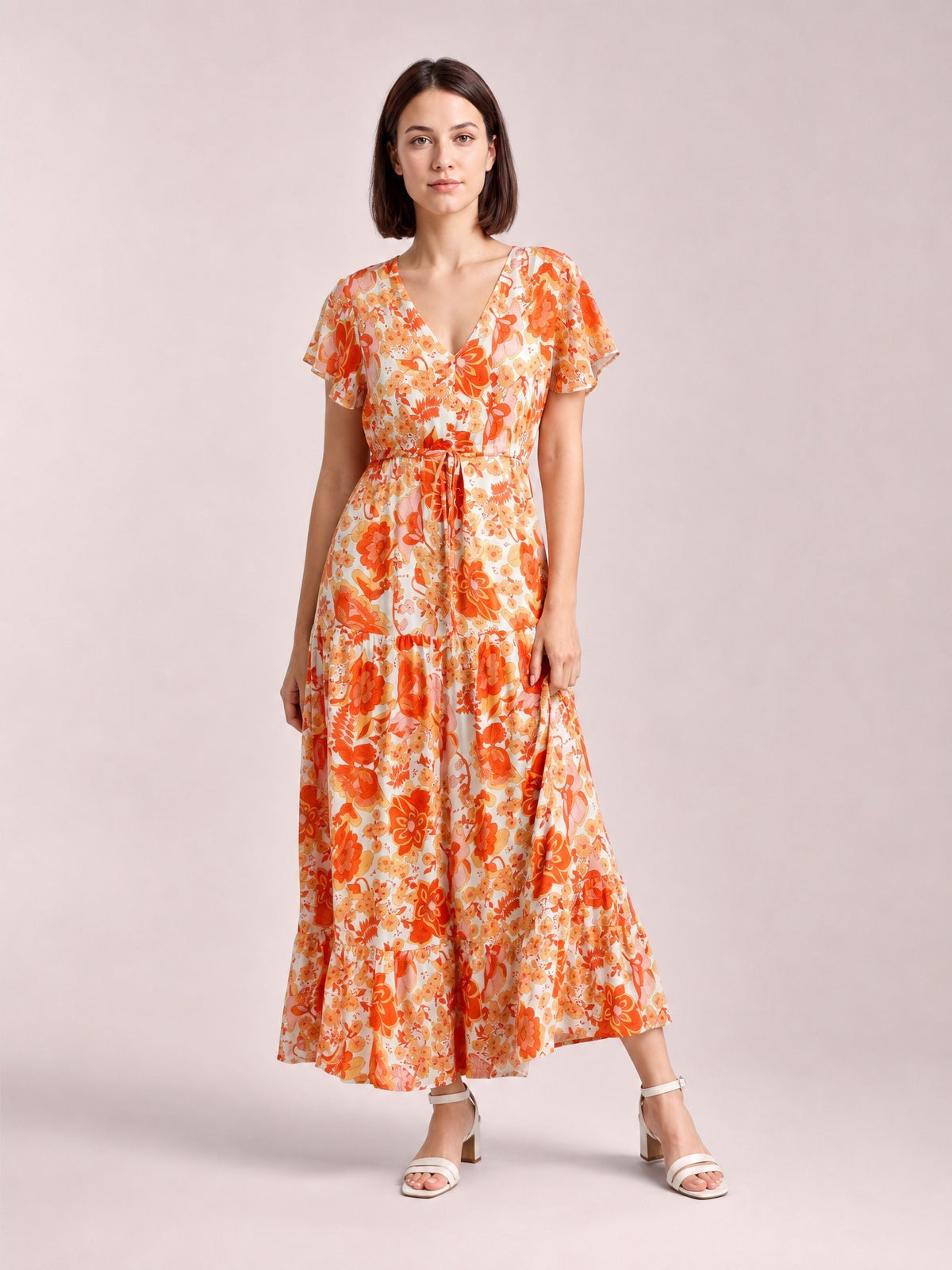 A model stands with one leg angled in an orange-and-cream floral midi dress featuring a V-neck, flutter sleeves, drawstring waist, and tiered skirt.