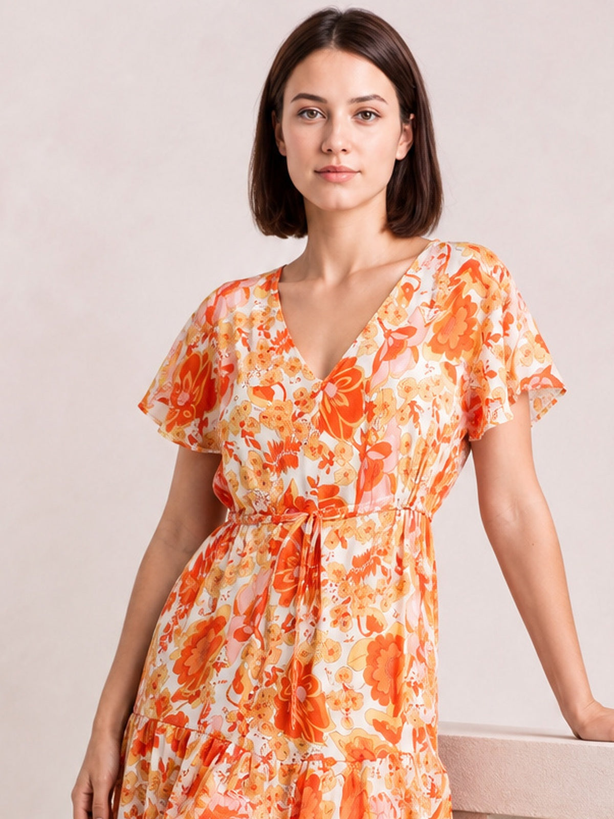 A model wears an orange and cream floral drawstring-waist tiered midi dress with a V-neckline and flutter sleeves, posing with one hand on a ledge.