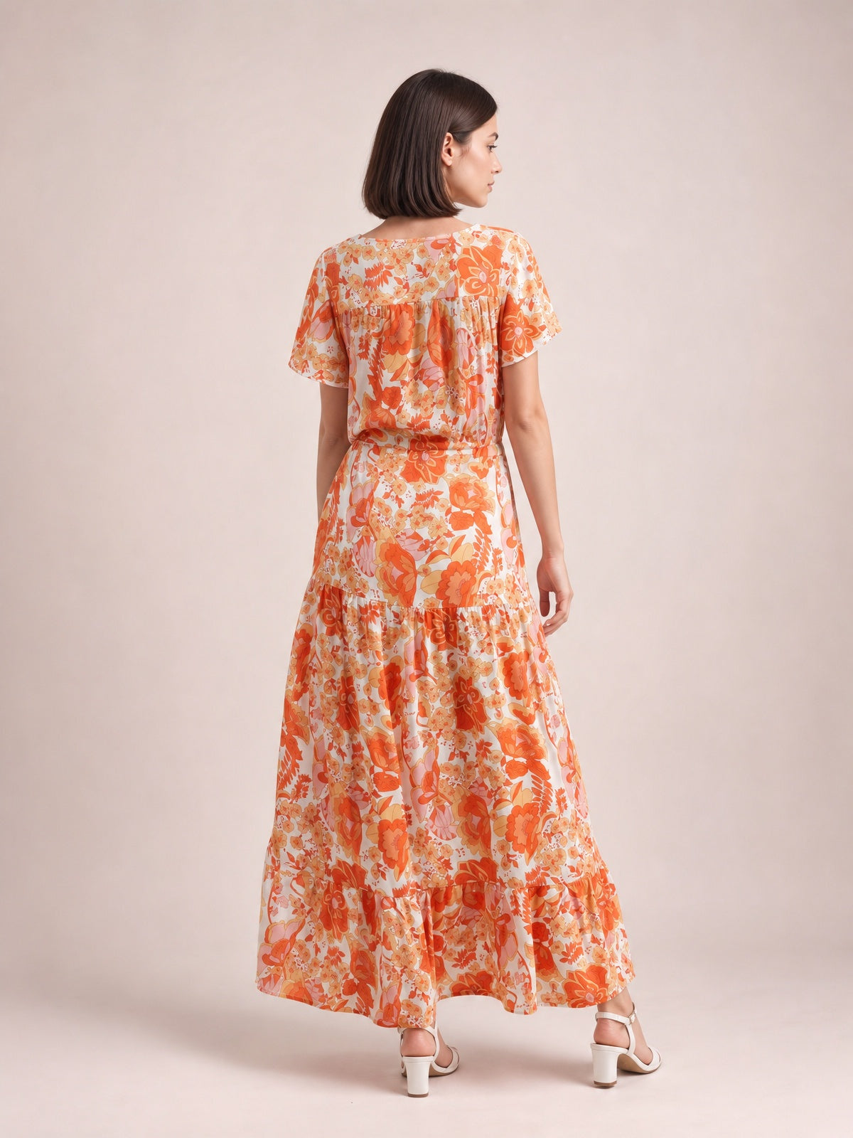 A model stands turned away in an orange floral midi dress with short flutter sleeves, a drawstring waist, and a flowing tiered skirt.