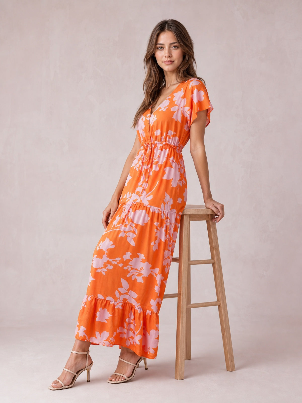 Model poses beside a stool in a bright orange floral midi dress with a V-neck, flutter sleeves, buttoned front, drawstring waist, and tiered skirt.