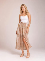 A model poses in a flowing beige floral maxi skirt with a smocked waist, buttoned front, and high slit creating an airy bohemian silhouette.