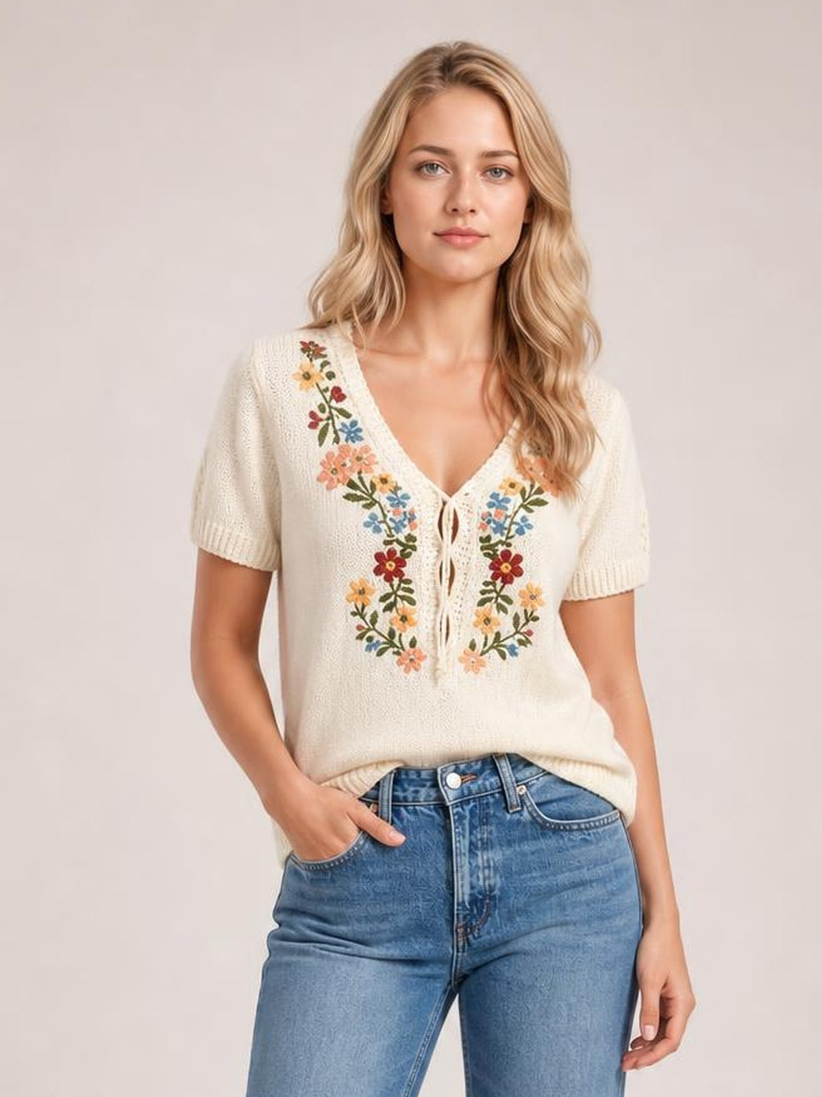 A woman poses with one hand in her jeans pocket, wearing a cream short sleeve sweater with a V-neck, tie front, and colorful floral embroidery.