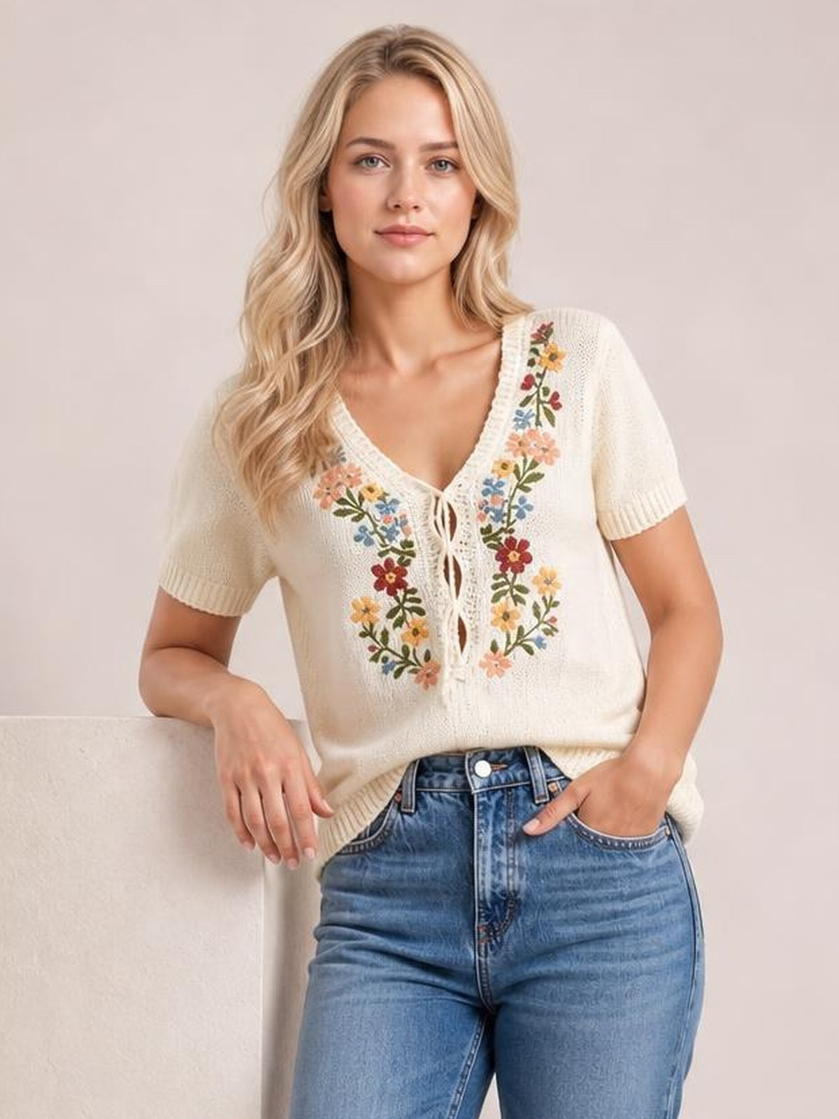 A model wears an ivory floral embroidered short sleeve sweater with a V-neck, lace-up front, ribbed cuffs and hem, and a relaxed silhouette.