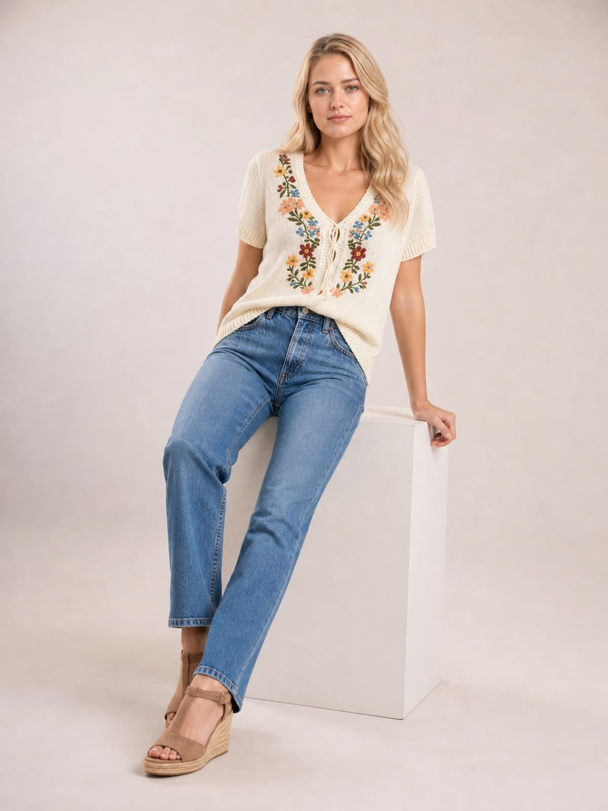 Model sits on a cube wearing a cream floral embroidered short-sleeve sweater with a V-neck, lace trim, tie front, and relaxed silhouette.