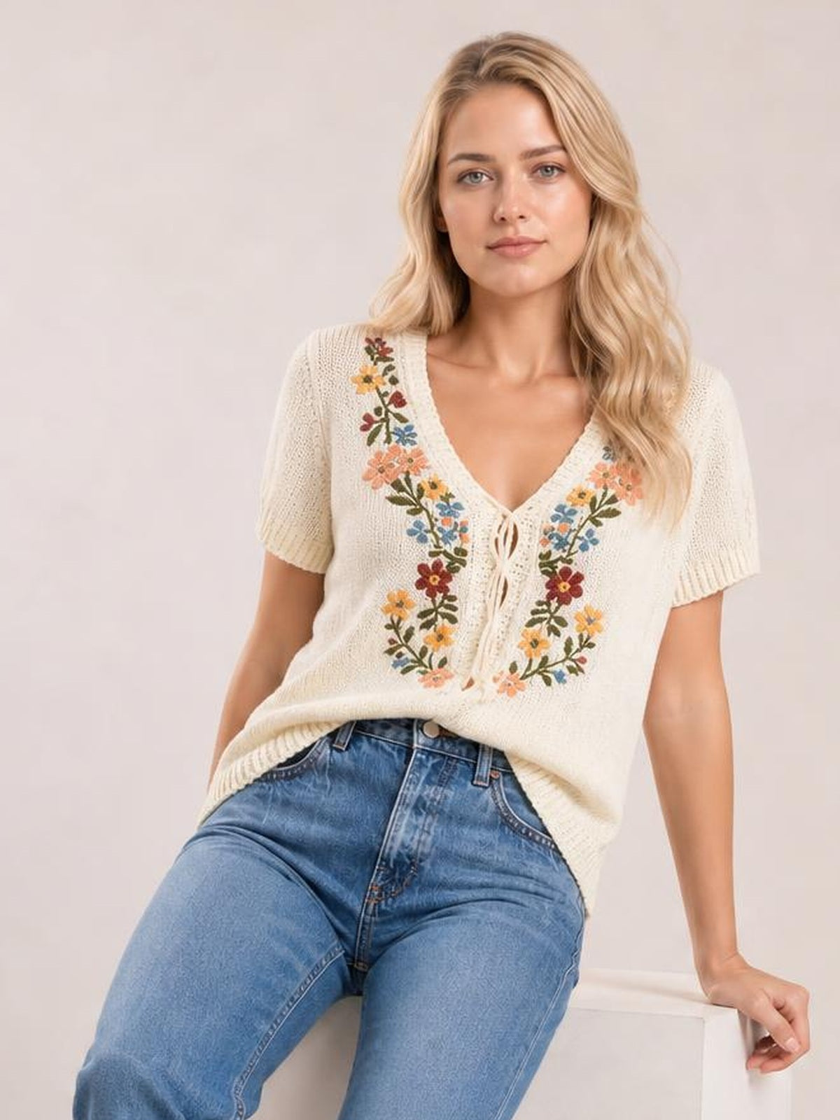 Model seated in a cream short-sleeve sweater with a V-neck lace-up front, colorful floral embroidery, ribbed cuffs, and a relaxed silhouette.