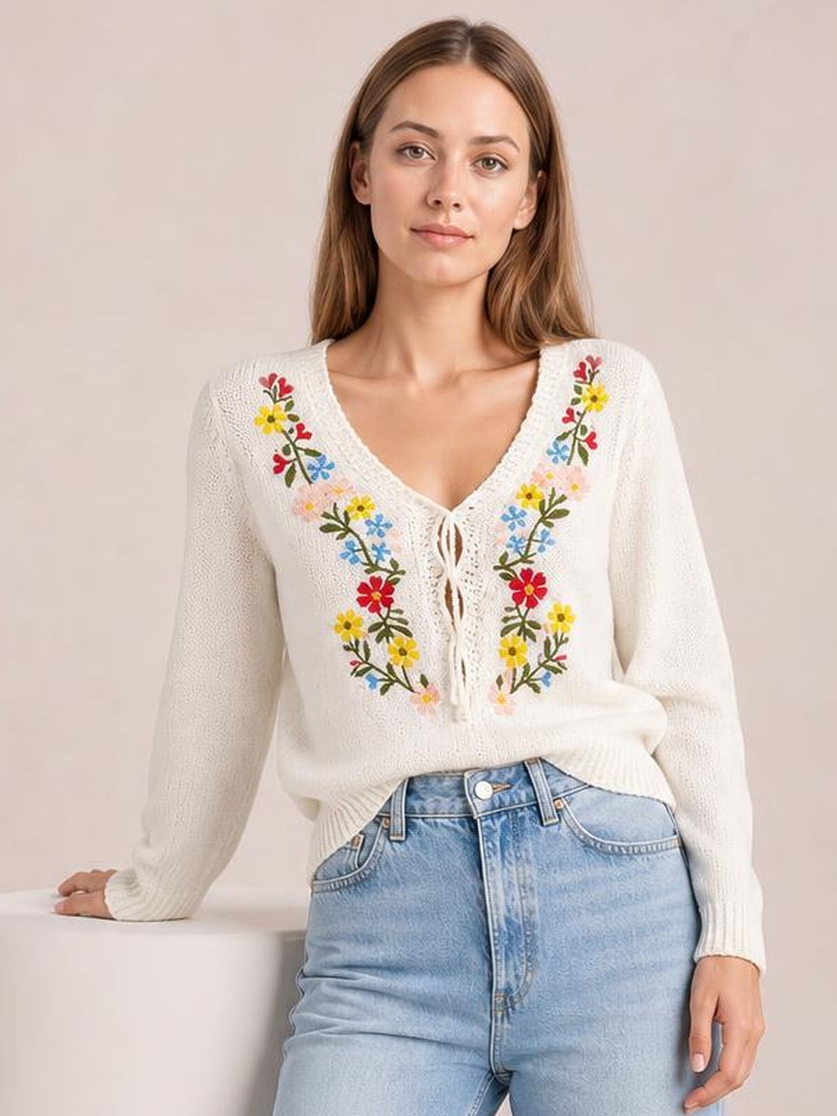 Model wearing a white floral embroidered V-neck sweater with long sleeves, a relaxed silhouette, and tied front detail, posing with one hand on a surface.