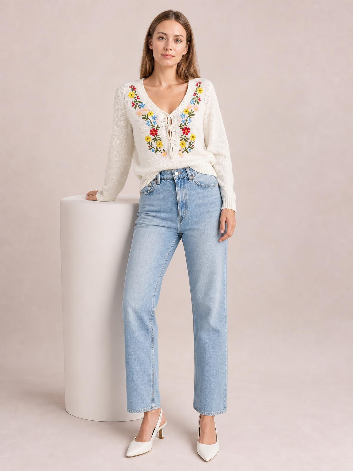 Model poses beside a pedestal wearing a white floral embroidered V-neck sweater with long sleeves, a relaxed silhouette, and a tied front detail.