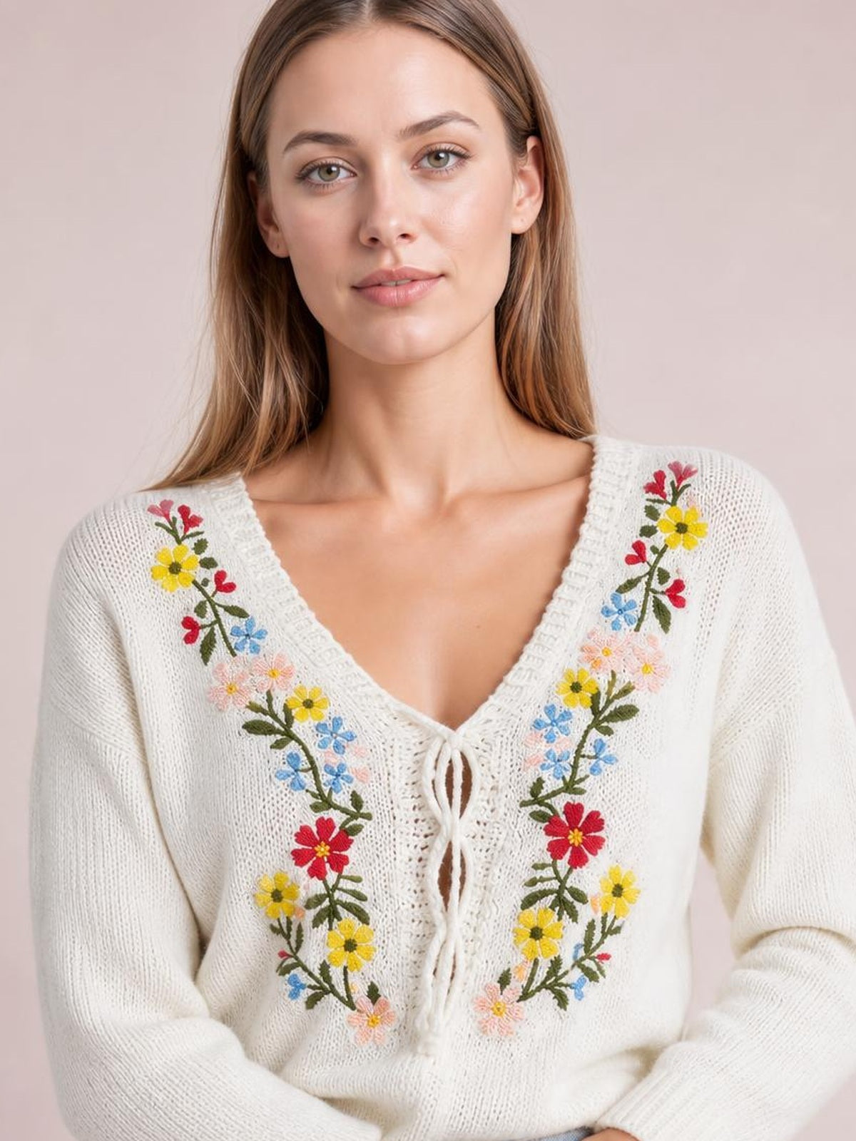 Model wears a white V-neck sweater with colorful floral embroidery, a tied front, long sleeves, and a relaxed knit silhouette while posing with hands folded.
