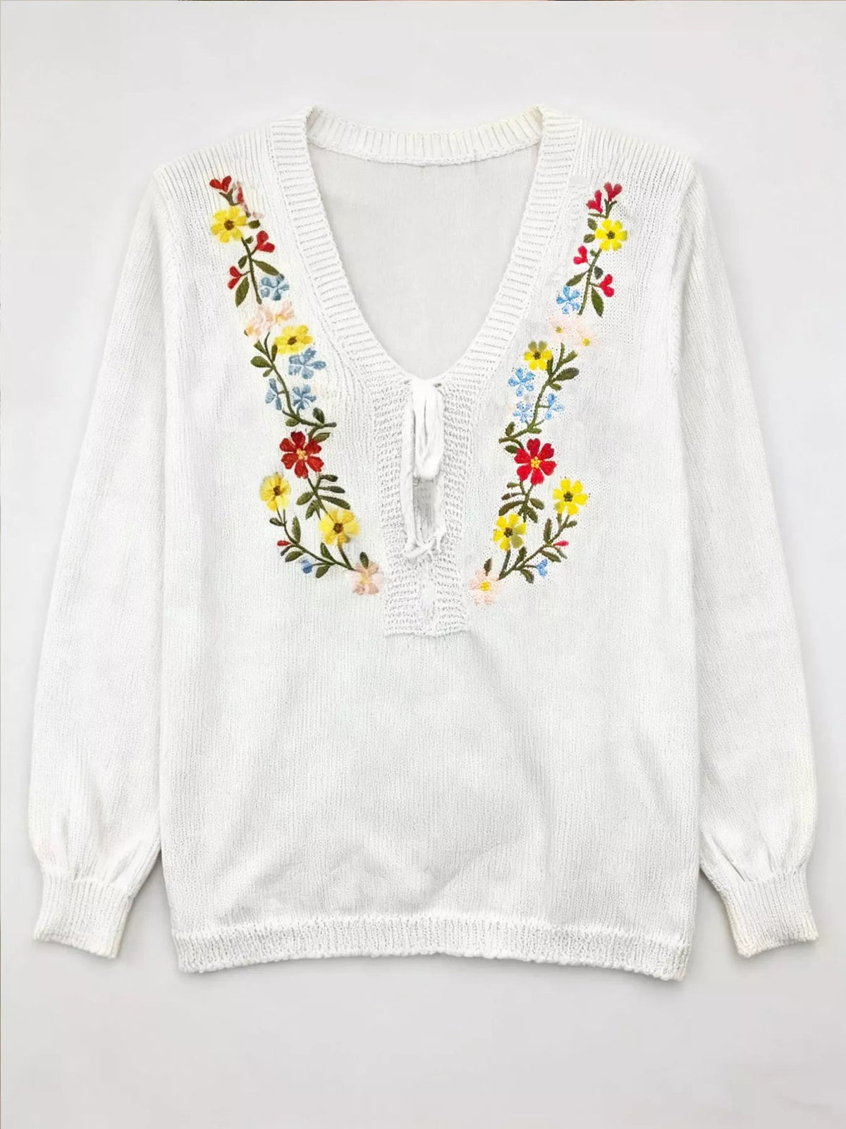 White knit V-neck sweater with long sleeves, ribbed trim, a relaxed silhouette, and colorful floral embroidery framing the neckline and front tie.