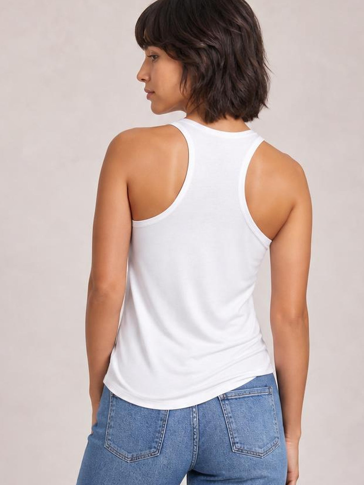 A model wears a white sleeveless tank with a high round neckline, racerback construction, smooth silhouette, and curved regular-length hem, shown from behind.