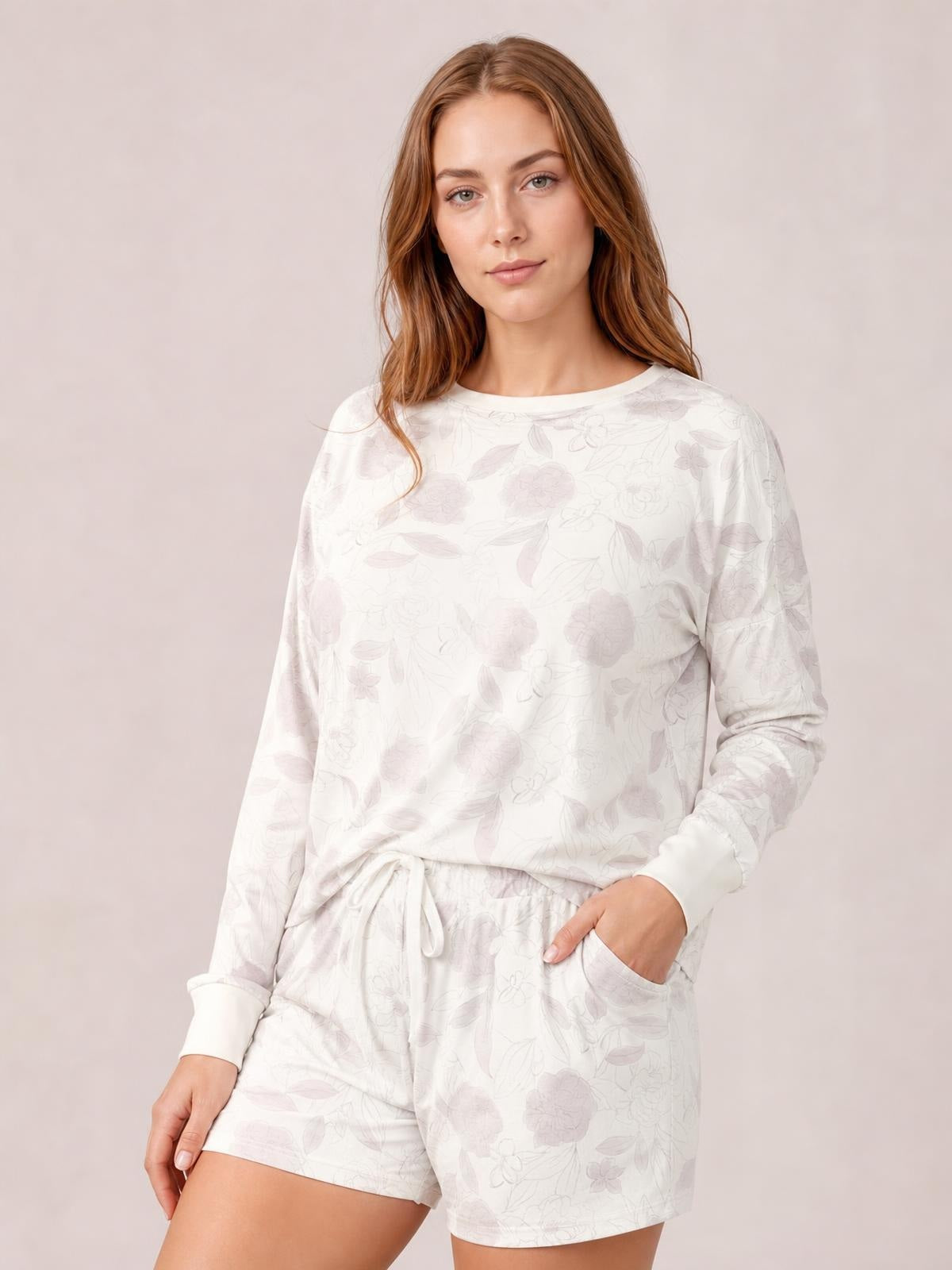 A model poses in a soft white floral loungewear set with a round-neck long-sleeve top, dropped shoulders, drawstring shorts, and one hand in a pocket.