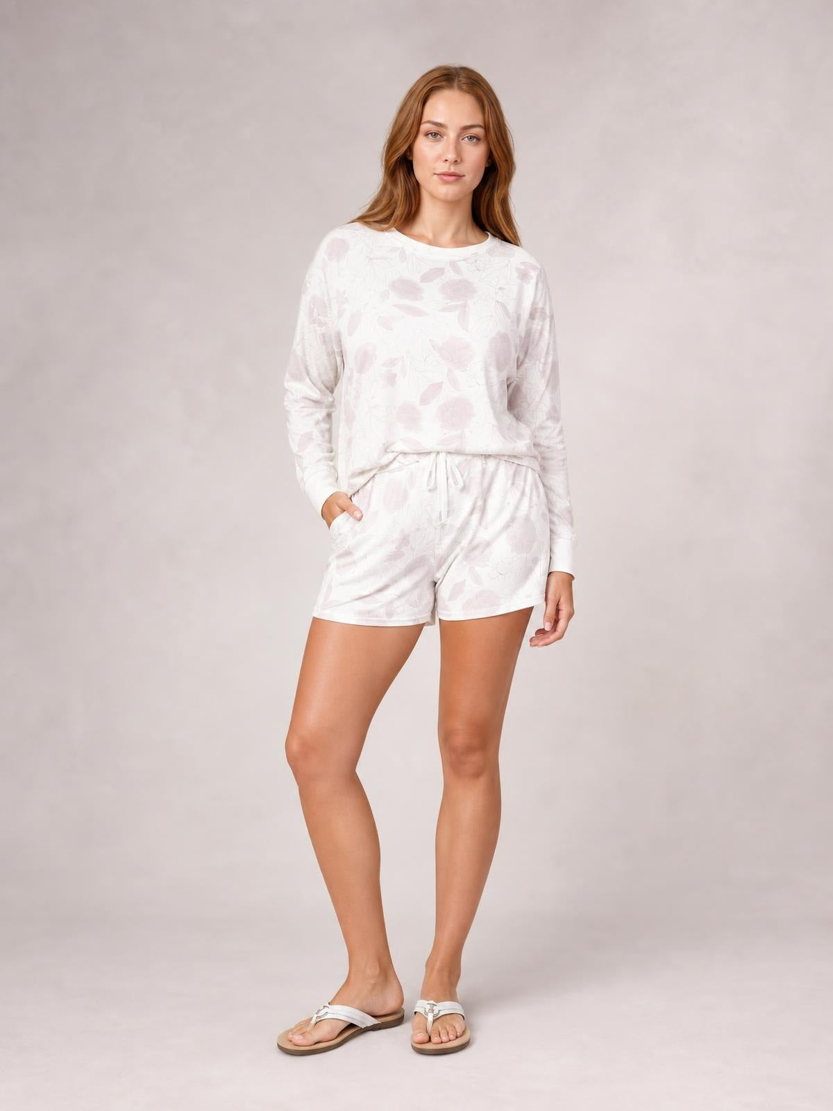 Model wears a pale floral loungewear set with a relaxed long-sleeve round-neck top and drawstring shorts, standing with one hand in a pocket.