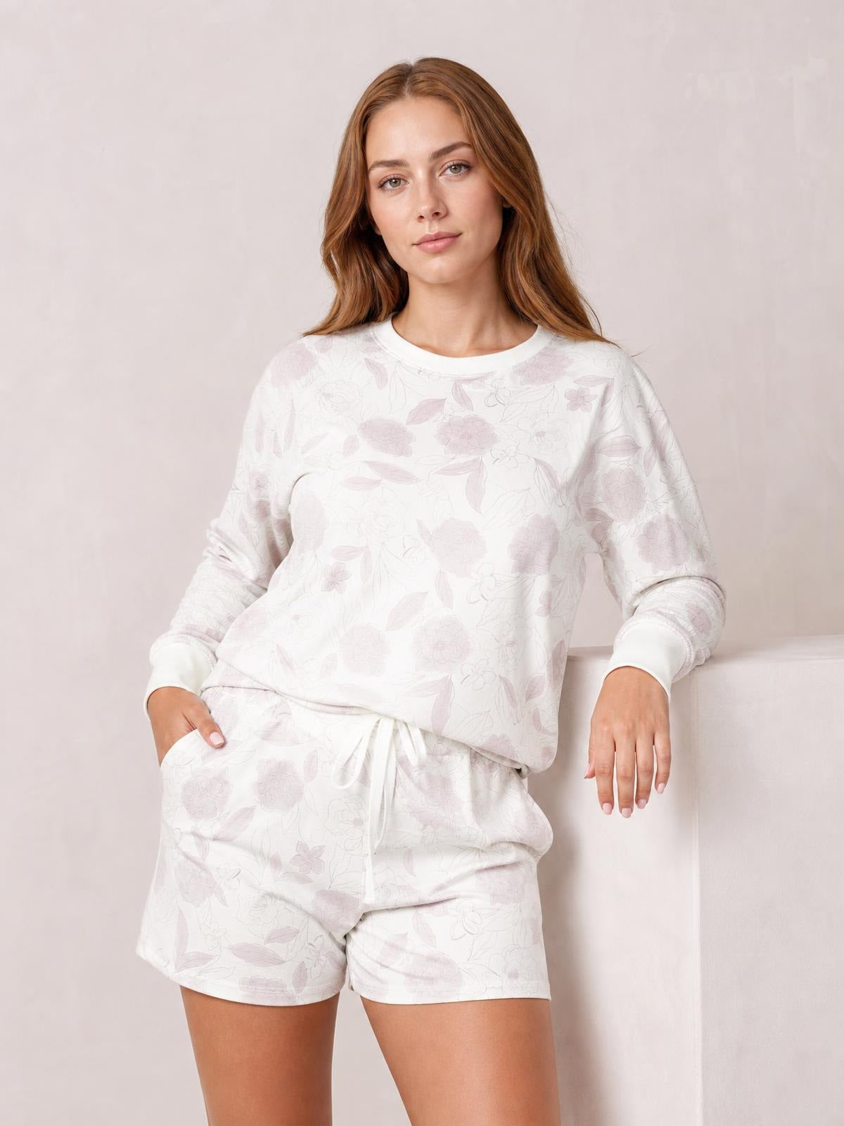 A model leans against a wall wearing a white floral long-sleeve top with a round neck and relaxed silhouette, paired with matching drawstring shorts.