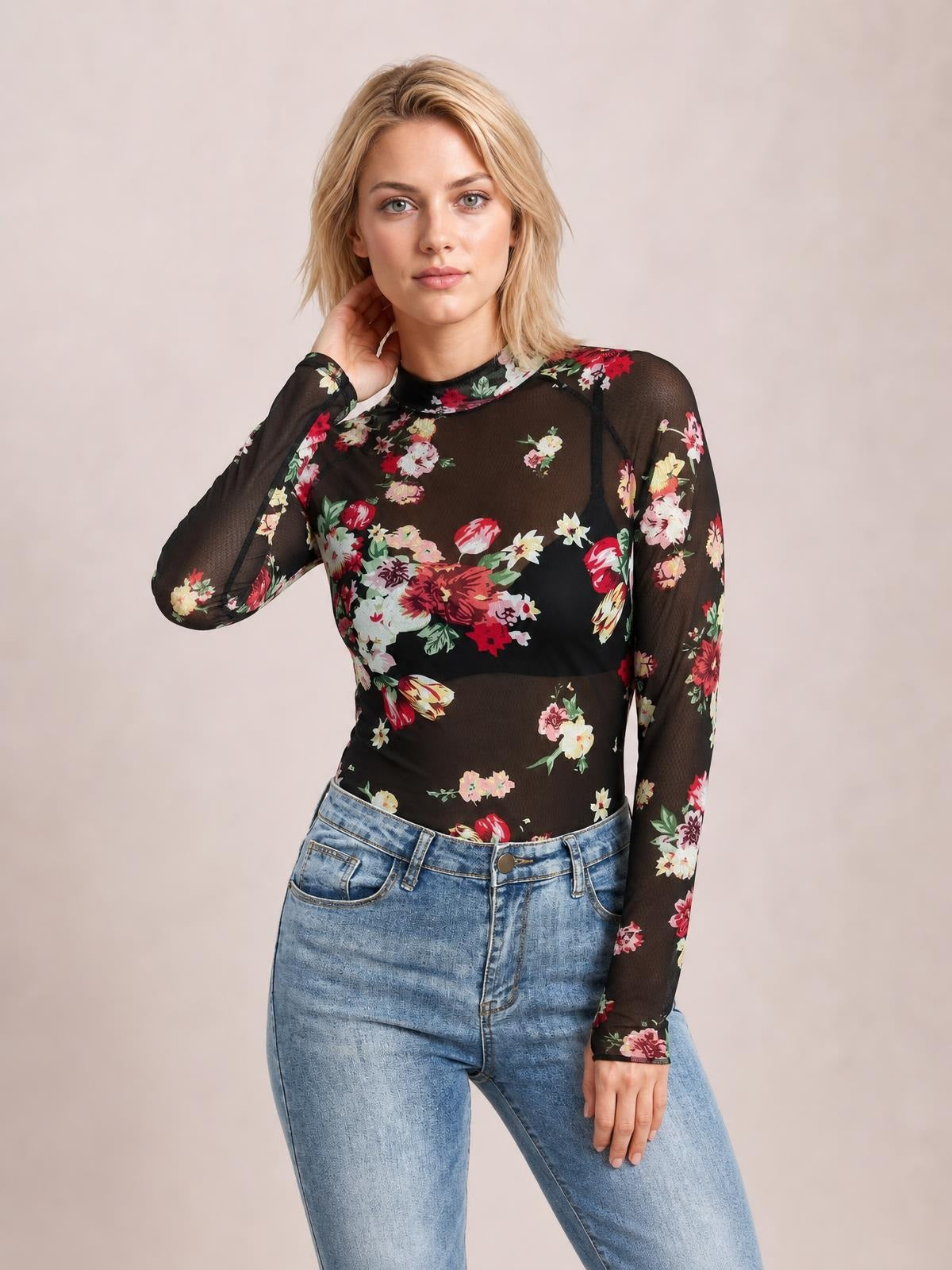 Model poses with one hand at her neck wearing a black floral mock neck long sleeve top with a fitted silhouette and sheer sleeves.