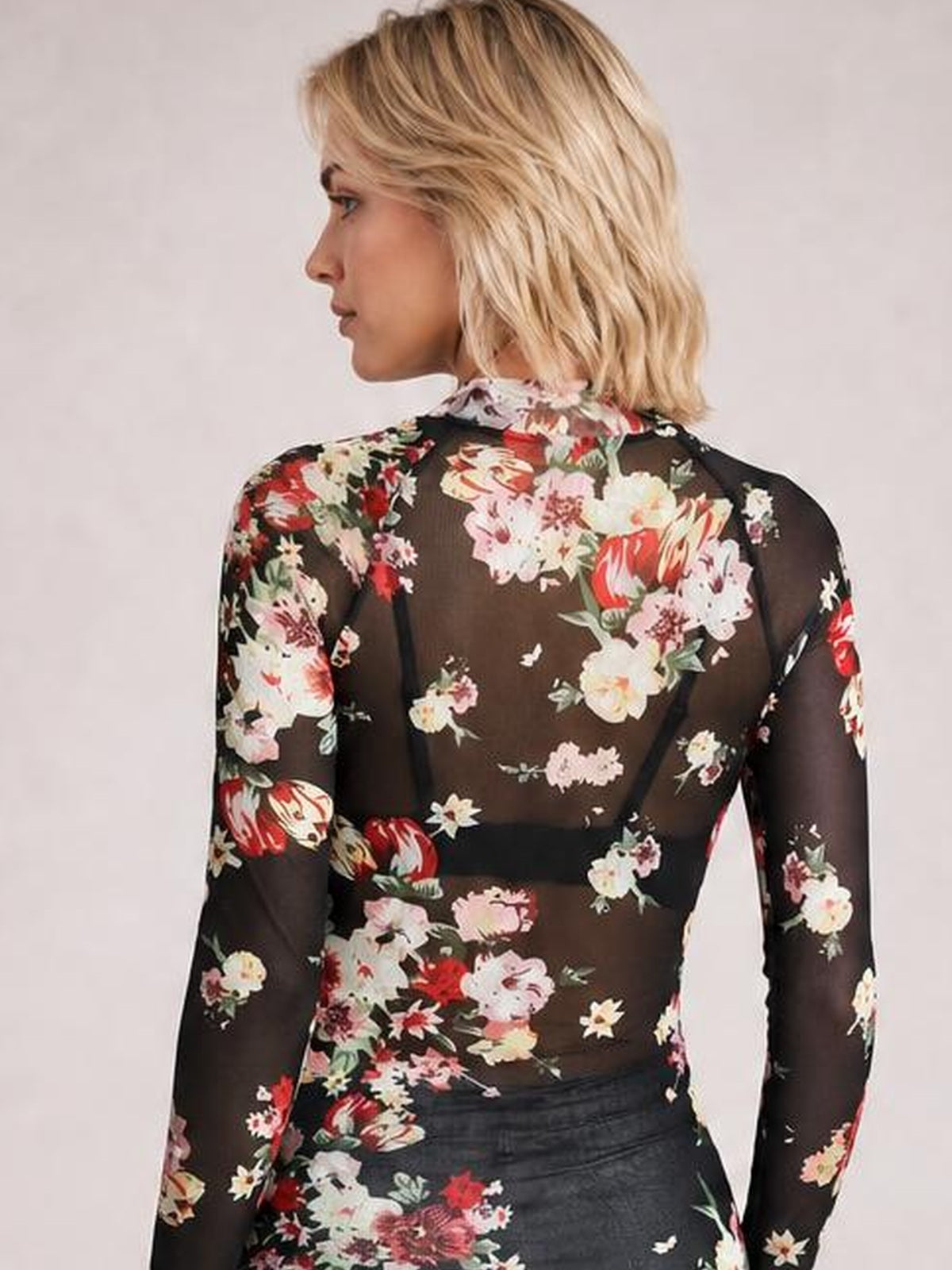 A model shown from behind wears a fitted black floral mock neck top with long sheer sleeves and colorful flower print throughout.