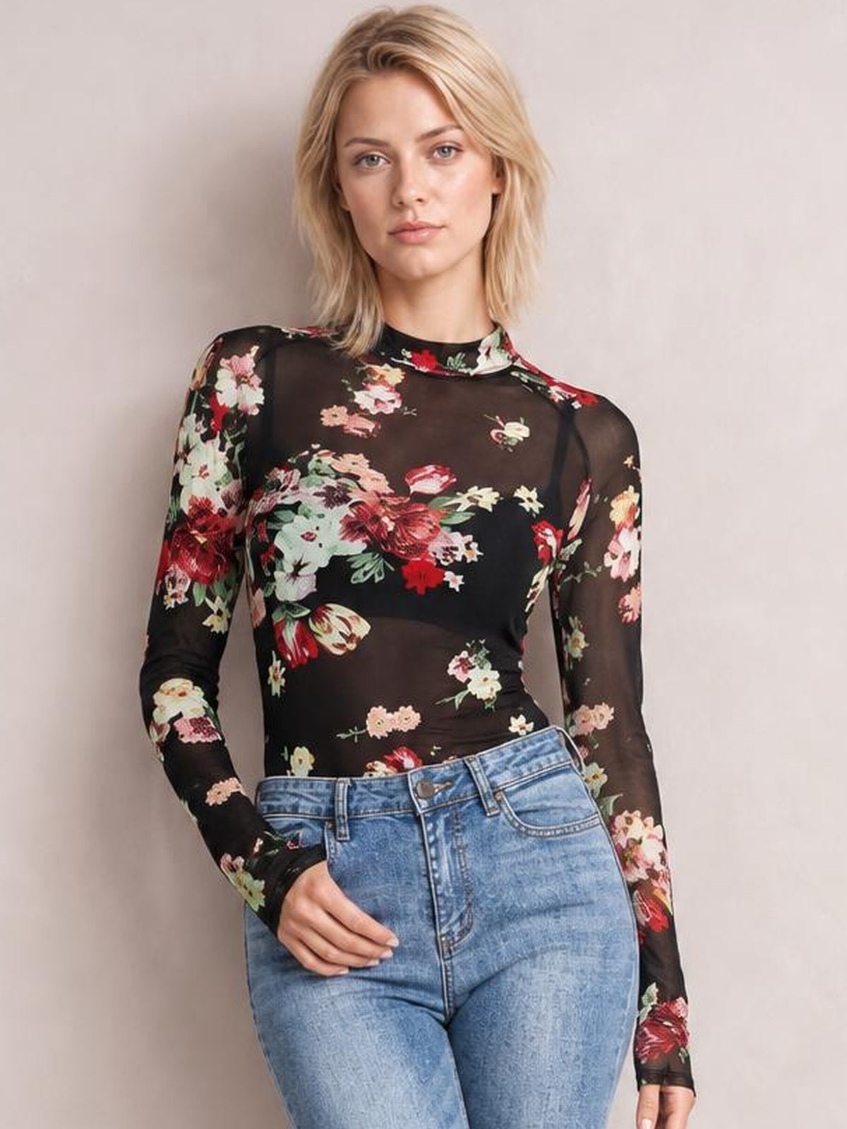 A model poses with one hand on her jeans, wearing a fitted black floral mock neck top with long sleeves and a softly sheer silhouette.