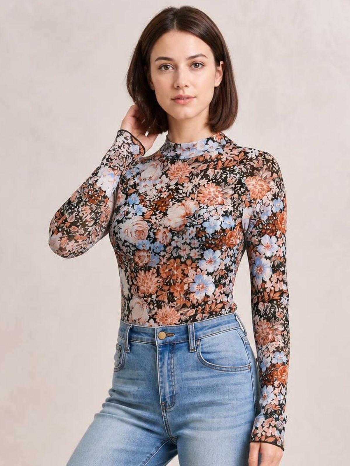 Model wearing a fitted floral mock neck long sleeve top with a black base and pink, blue, and orange flowers, posing with one hand near her hair.