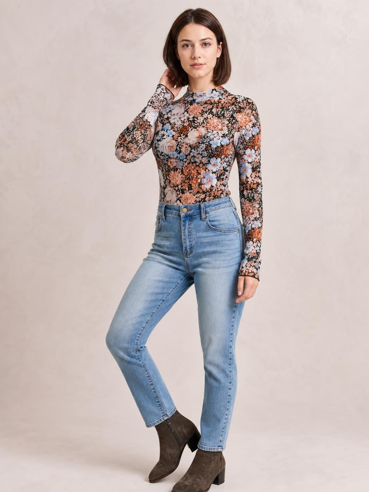Model poses in a fitted floral mock neck long sleeve top with a sheer black base and multicolor flowers, tucked into light-wash jeans.