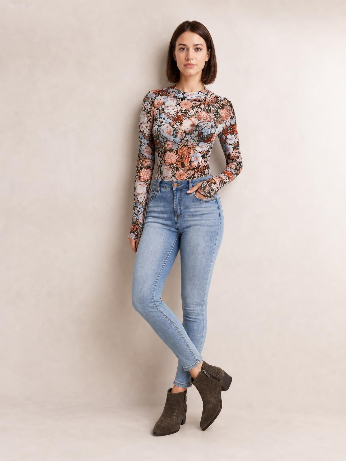 Model wears a fitted floral mock neck long sleeve top with a multicolor botanical print and smooth stretch silhouette, posing with one hand in her jeans pocket.