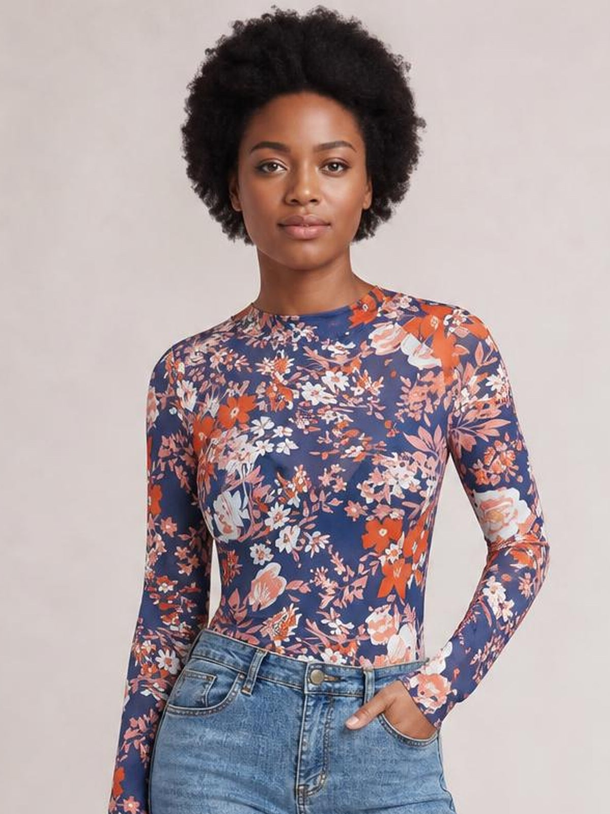 Model poses with one hand in her jeans pocket wearing a navy floral mock neck long sleeve top with a fitted silhouette and colorful flower print.
