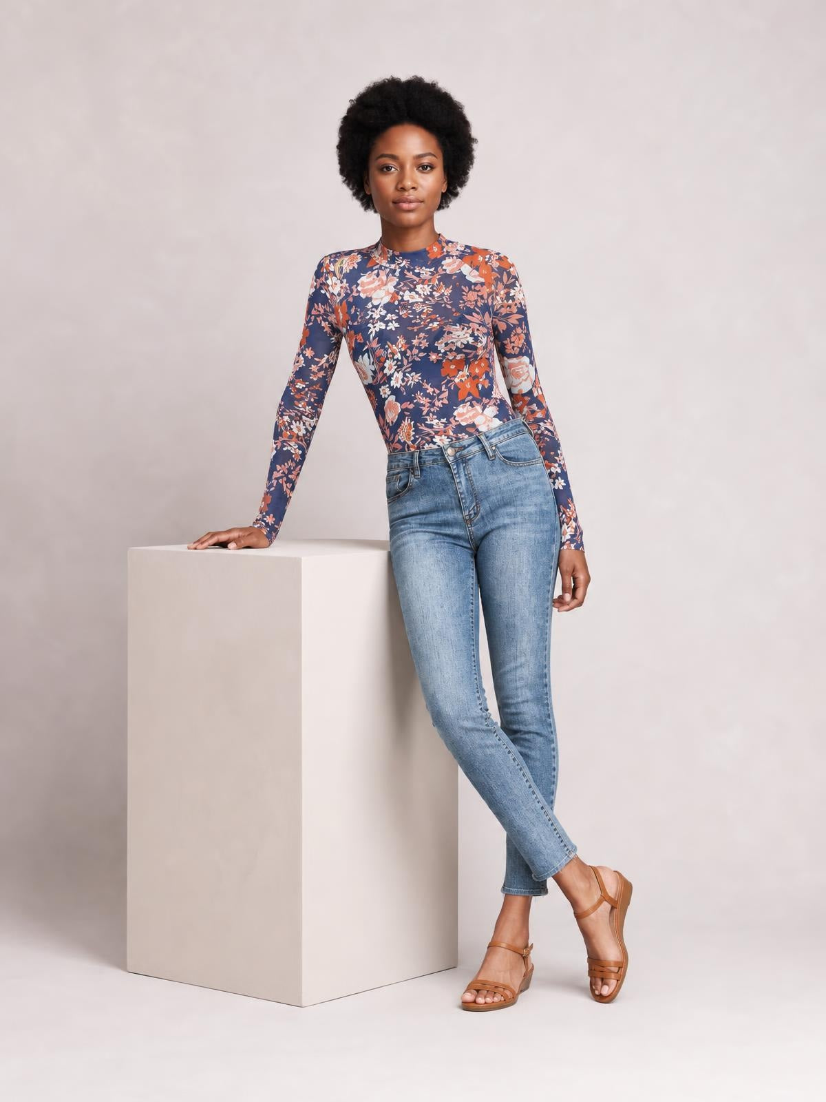 A model wears a fitted navy floral mock-neck top with long sleeves, leaning one hand on a block with her legs crossed.