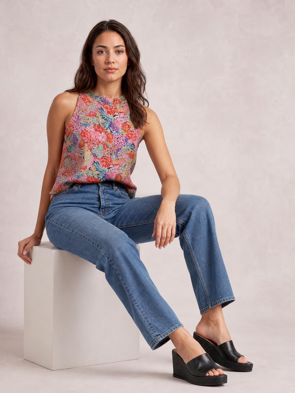 Model seated in a relaxed pose wears a colorful floral mock neck tank top with a sleeveless cut, regular length, and softly tailored silhouette.