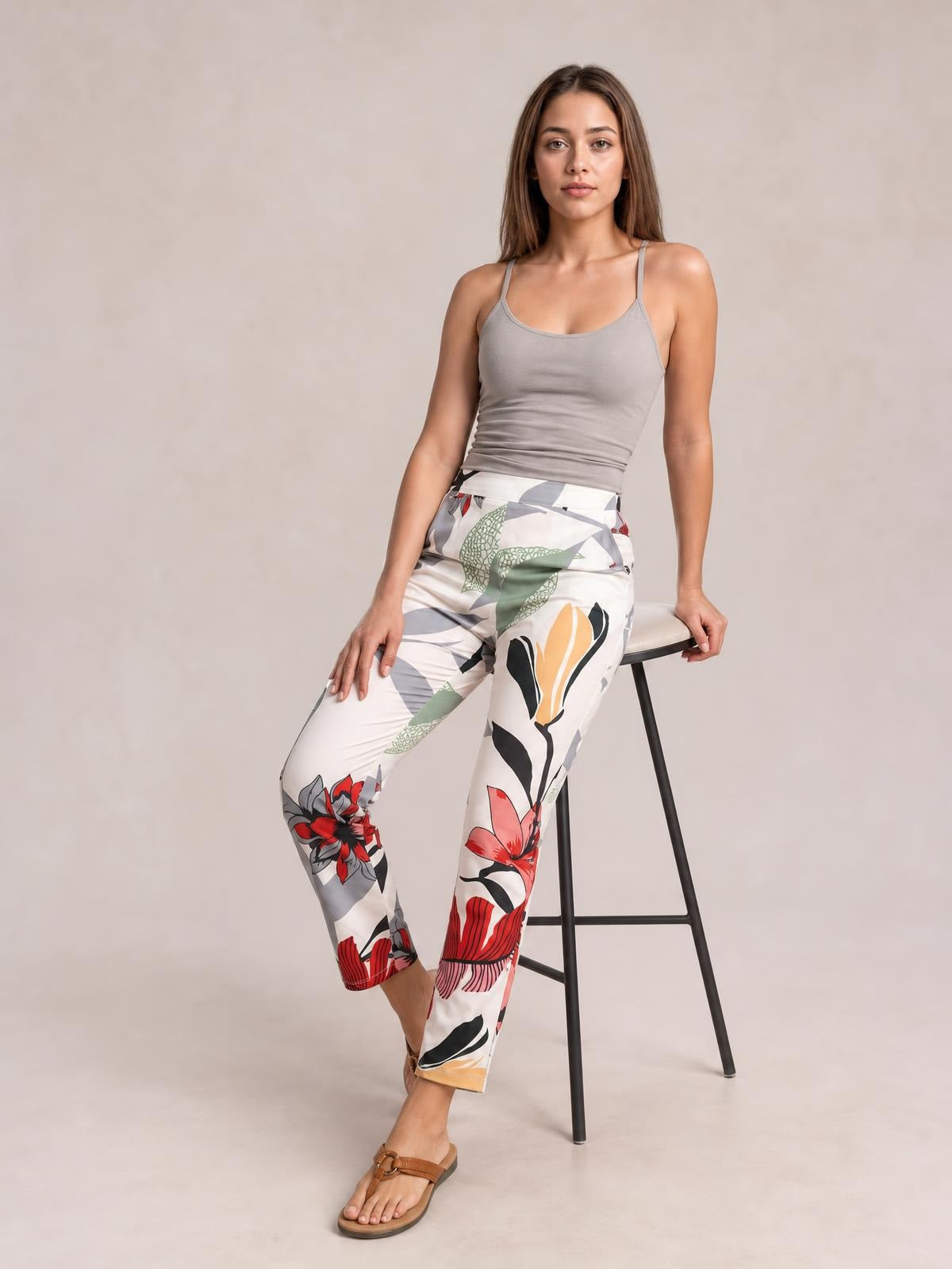 Model wears white floral print cropped pants with colorful botanical motifs, a fitted straight-leg silhouette, and pockets while posing beside a stool.