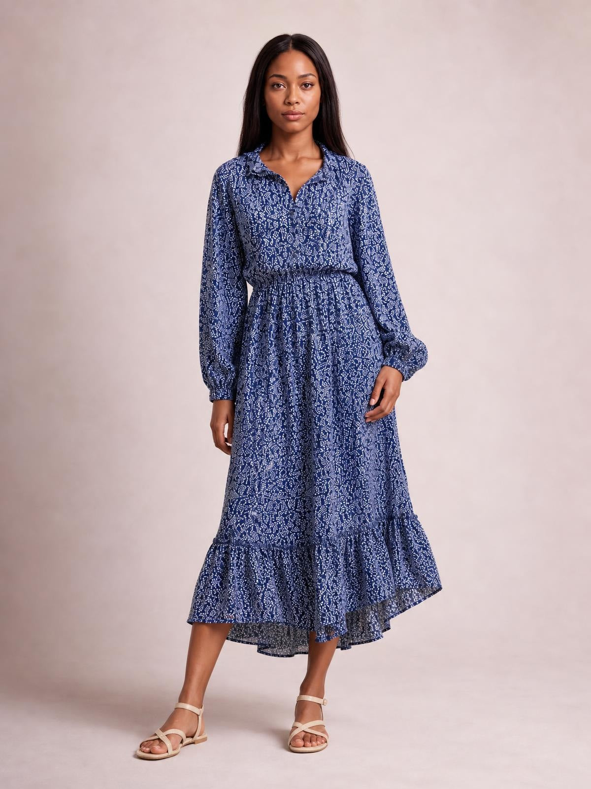 Model wears a blue floral print midi dress with long sleeves, a smocked waist, flowing silhouette, and tiered ruffle hem, standing with one hand at the skirt.
