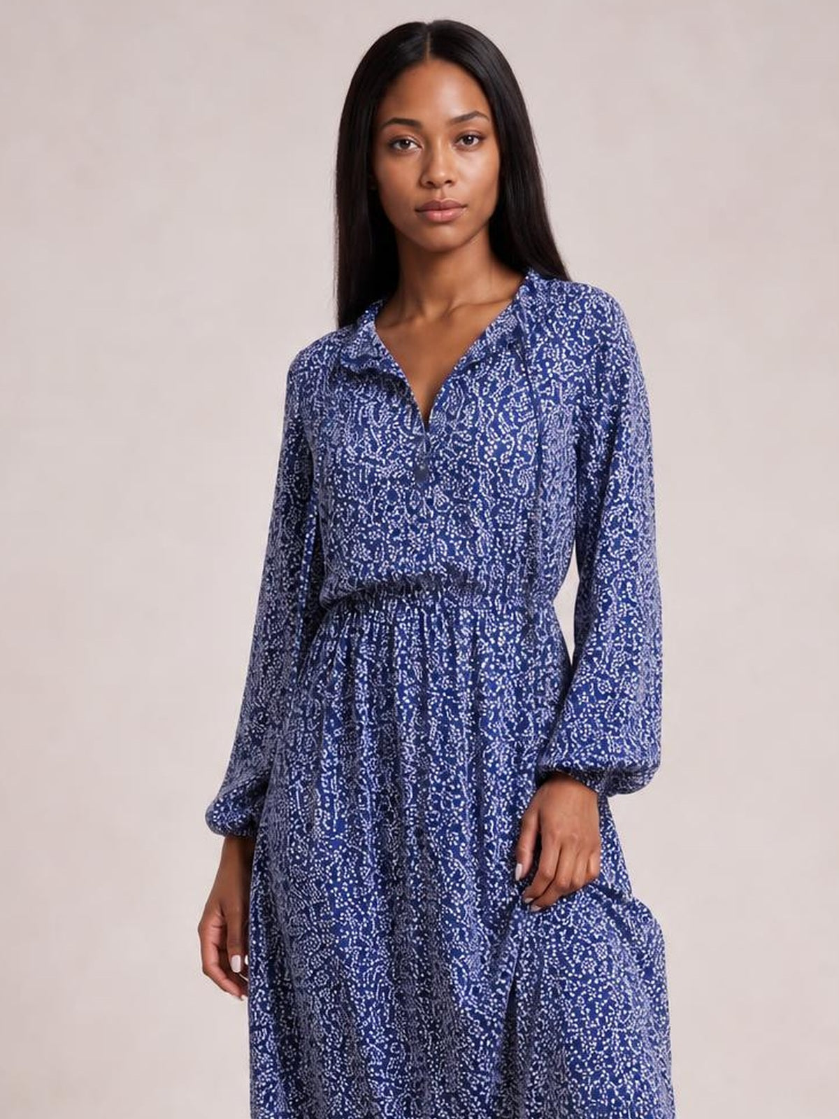 A model stands in a blue and white floral-print midi dress with long sleeves, a smocked waist, ruffled hem, and softly flared silhouette, holding the skirt.