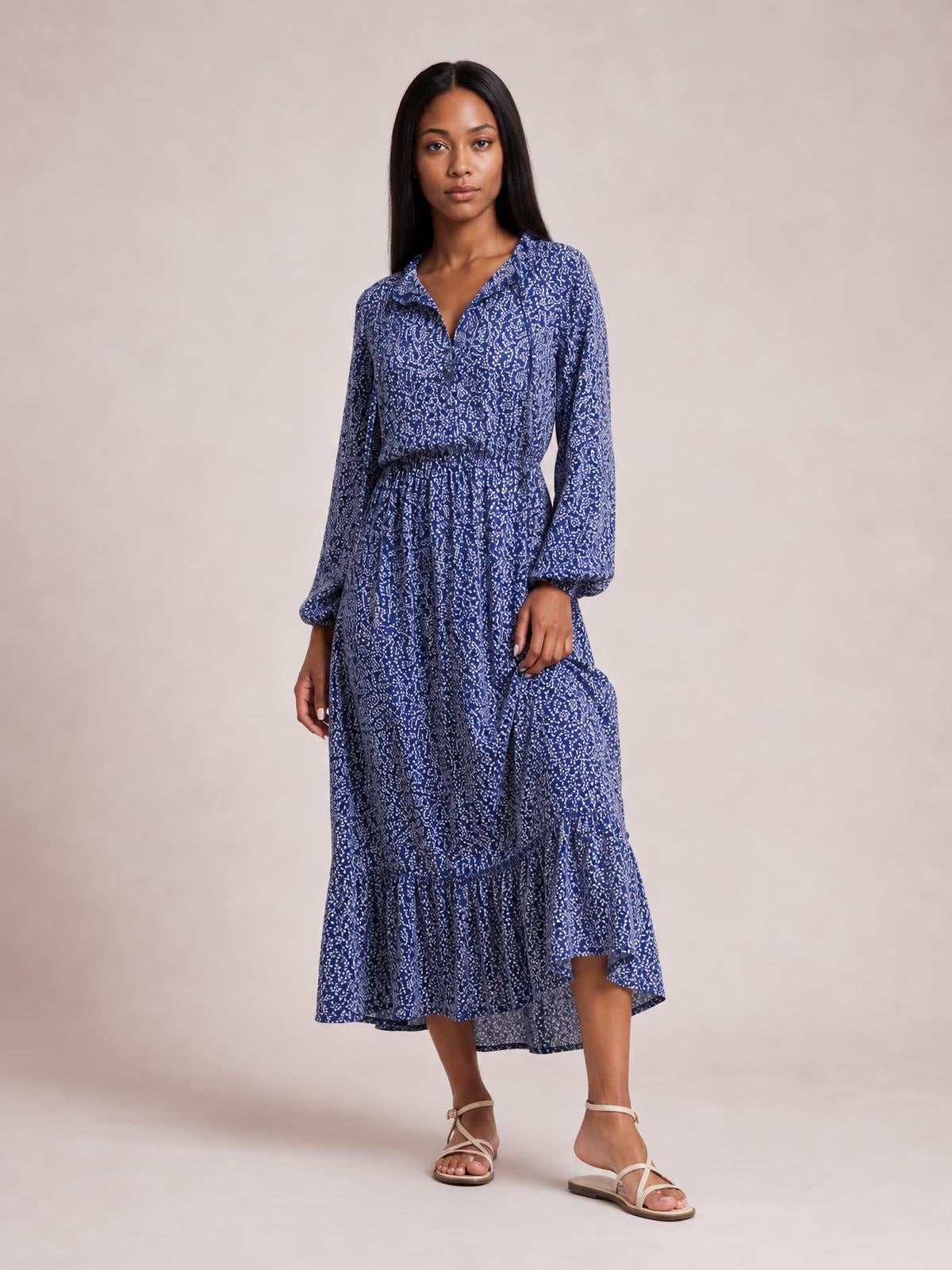 A model poses in a blue floral print long-sleeve midi dress with a smocked waist, flowing silhouette, and tiered ruffle hem.