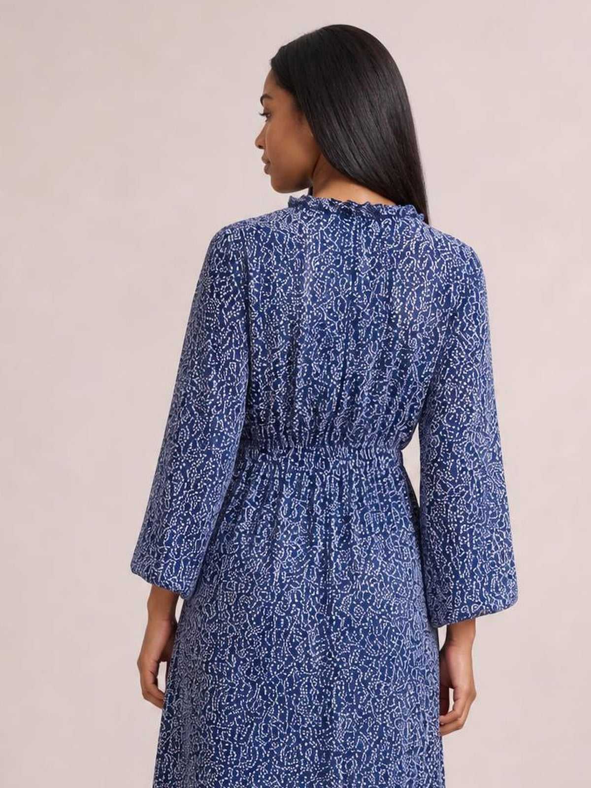 A model shows the back of a blue floral print midi dress with long sleeves, a ruffled neckline, smocked waist, and relaxed flowing silhouette.