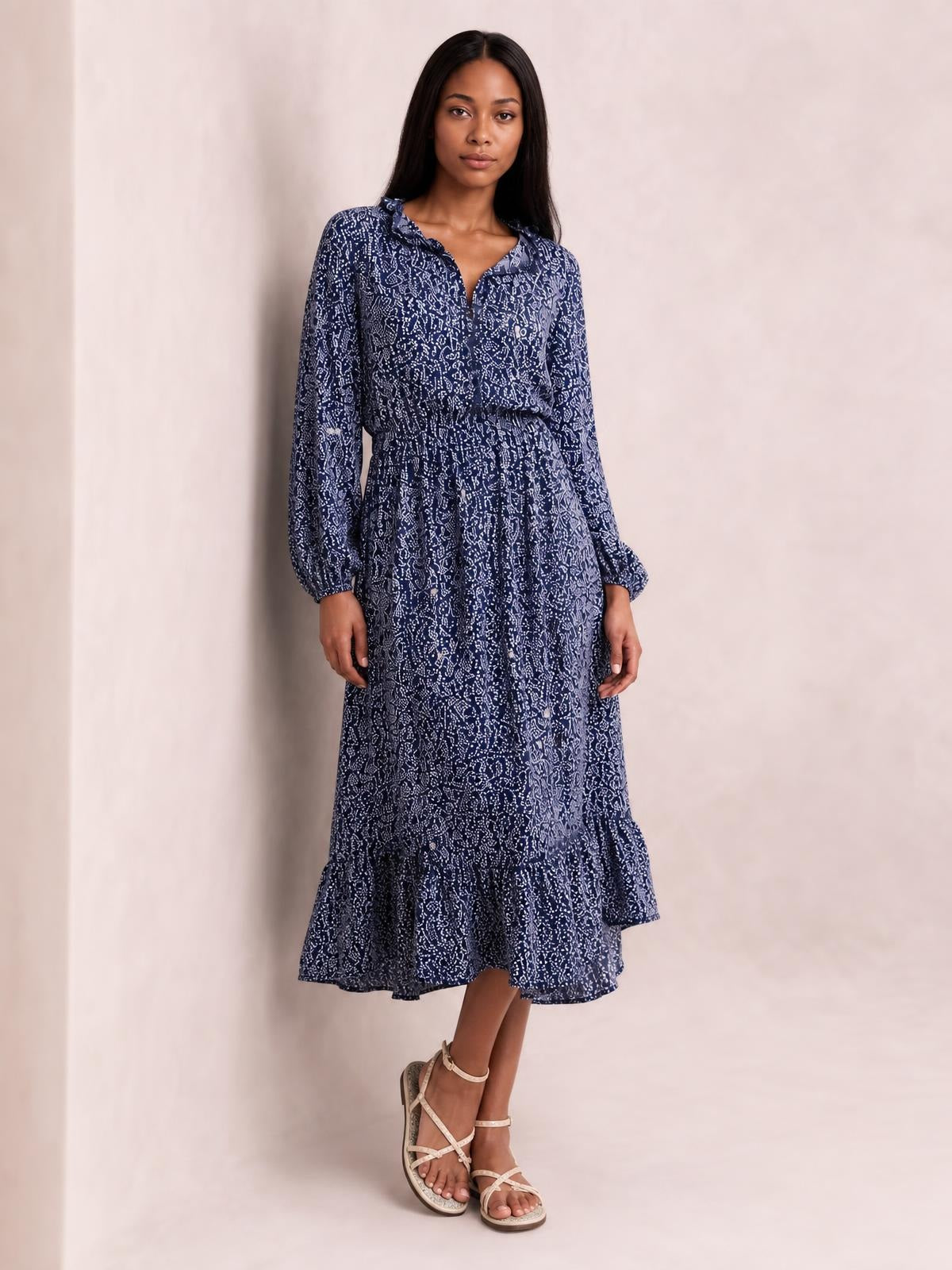 Model stands with crossed legs in a navy floral print midi dress featuring long sleeves, a ruffled neckline, smocked waist, button front, and tiered ruffle hem.