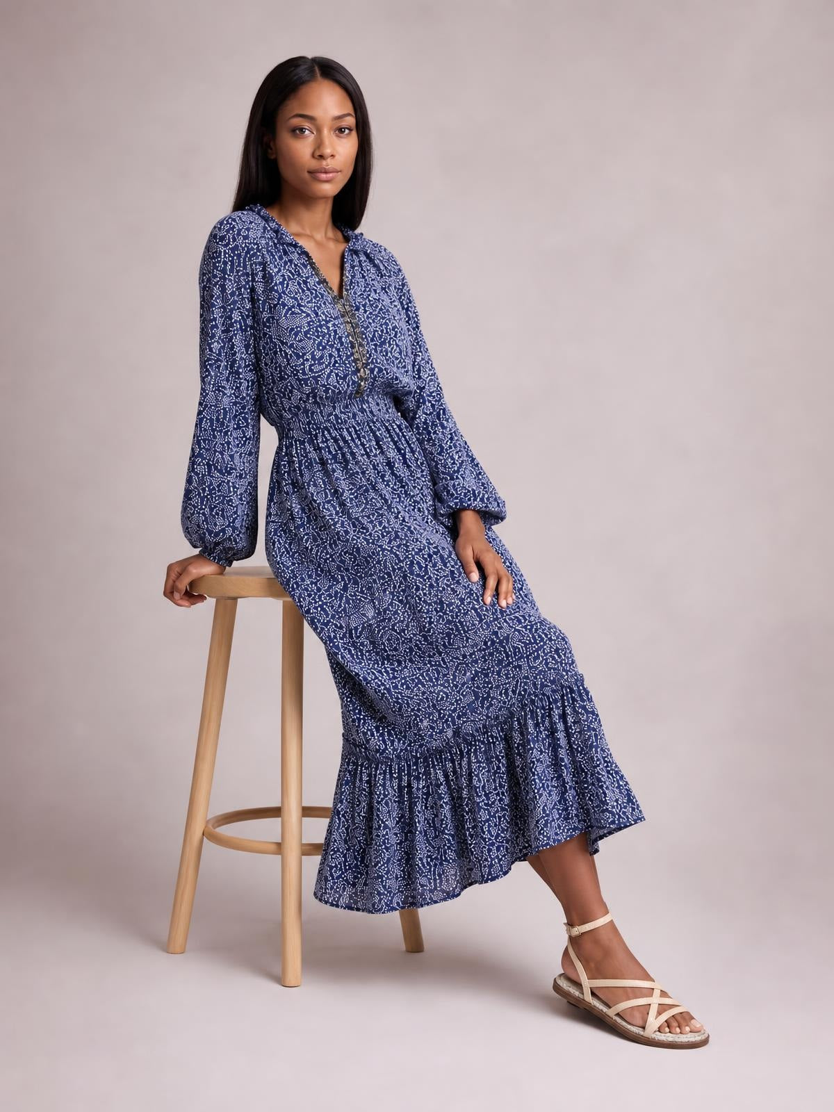 A seated model wears a navy floral print midi dress with long balloon sleeves, a smocked waist, flowing silhouette, and tiered ruffle hem.