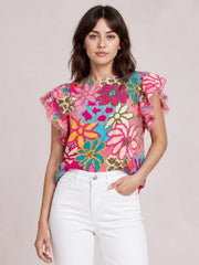 Model poses wearing a bright floral print round-neck blouse with a regular silhouette and short flutter sleeves featuring ruffled edges.