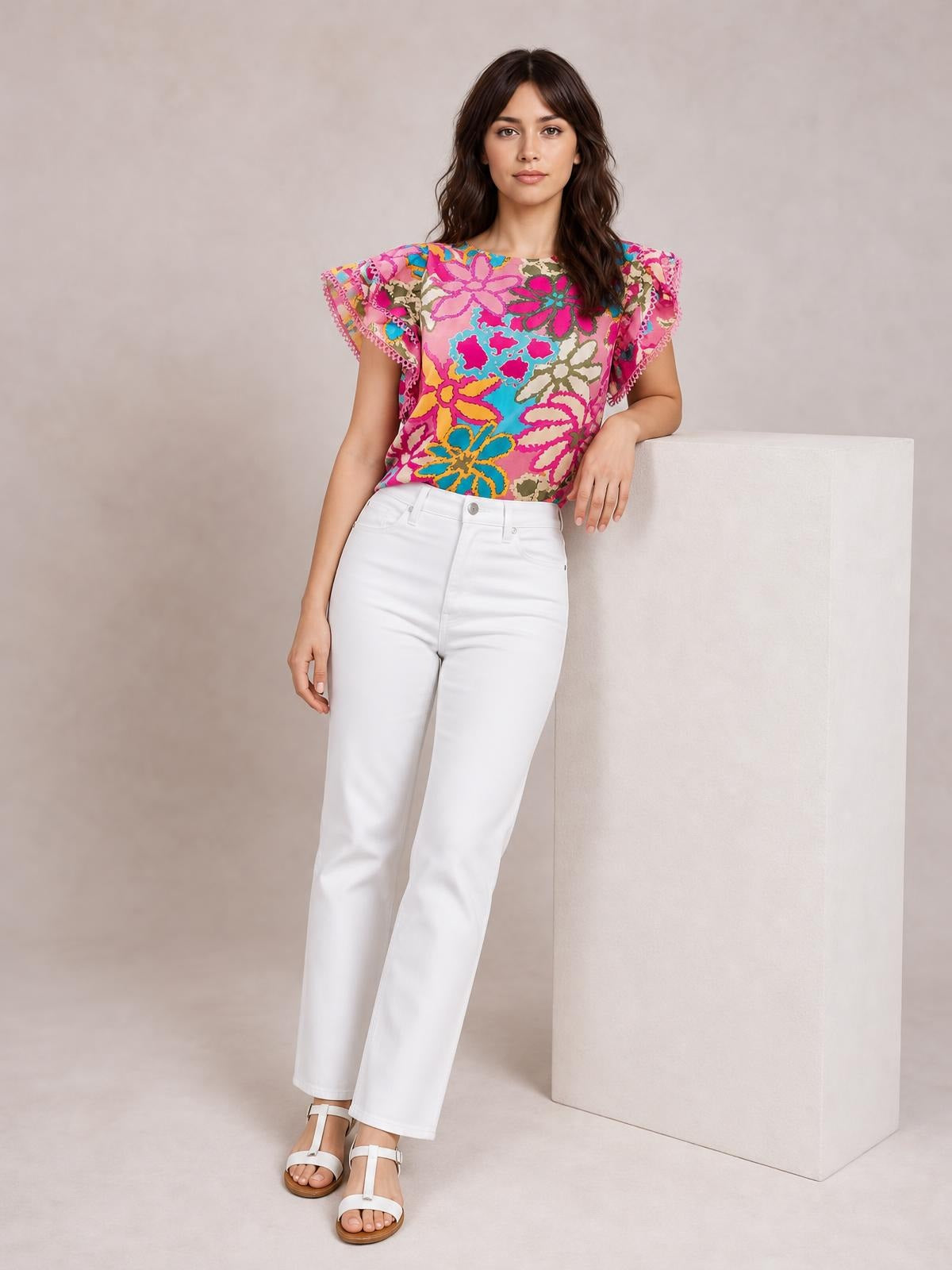 Model poses leaning on a pedestal in a vibrant floral print blouse with a round neck, flutter sleeves, and a regular silhouette.