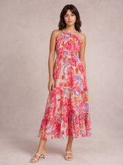 Model stands in a colorful floral print maxi dress with a smocked bodice, thin straps, defined waist, and flowing tiered skirt in pink, coral, orange, and lavender.